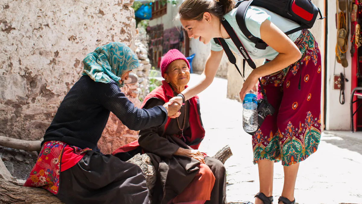 A student carrying a backpack and camera leans down to shake hands with an older woman seated on a log in a stone village lane, a second woman beside her