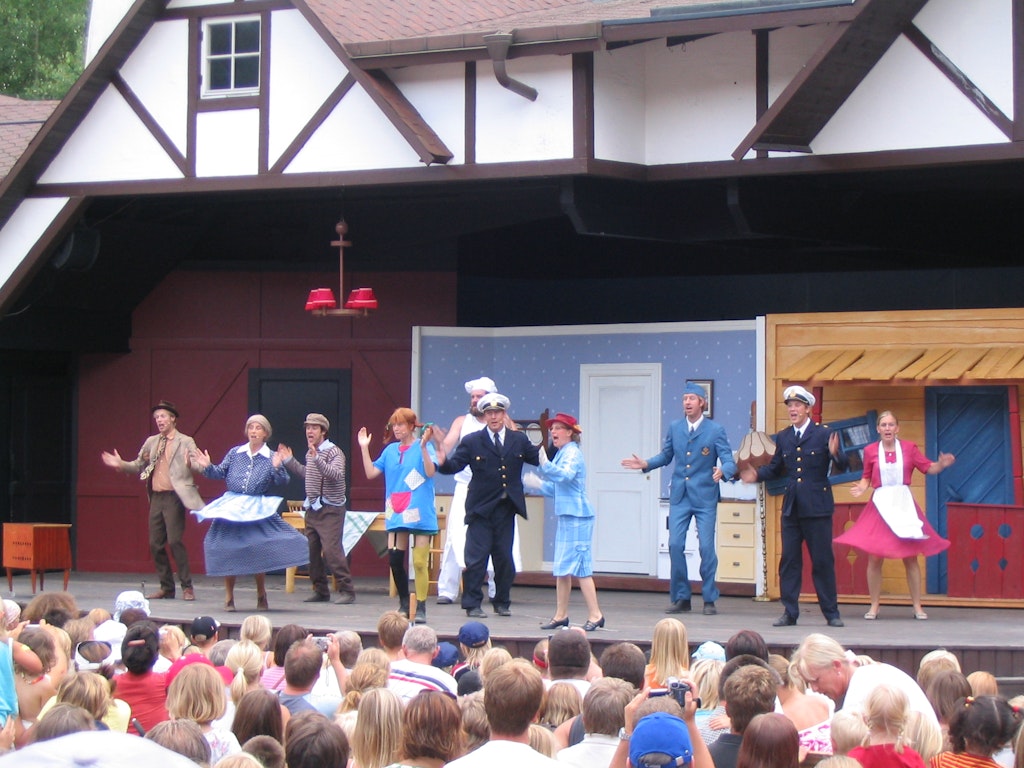 A lively outdoor stage performance with actors in colorful costumes portraying various characters, entertaining a large family audience.