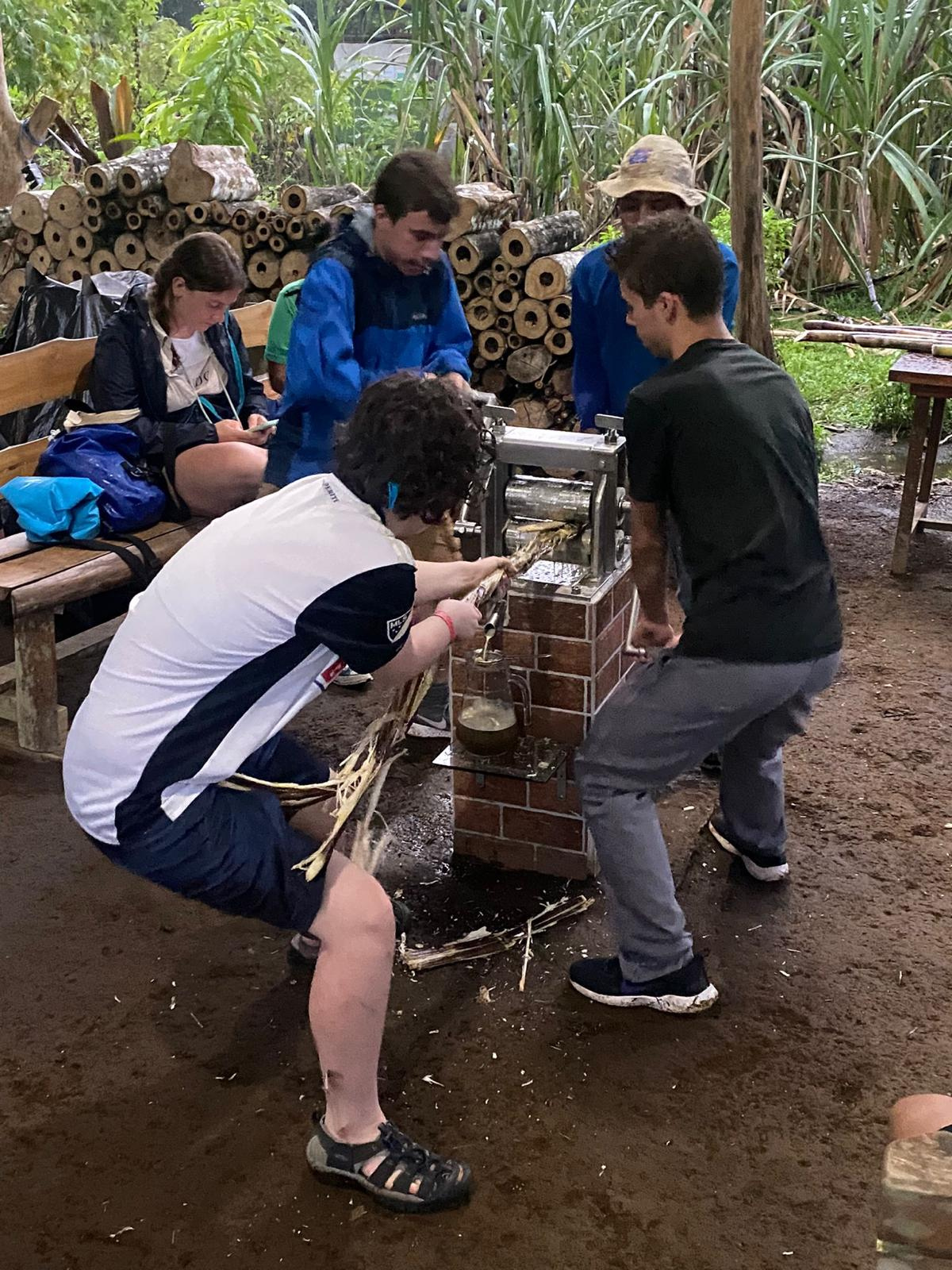 Students seen working on something together outside. One guy dragging some tree through a machine with the help of other guys.