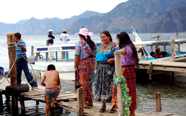 Women in embroidered blouses talking on a wooden lake dock beside moored passenger launches, with a volcanic ridge across the water.