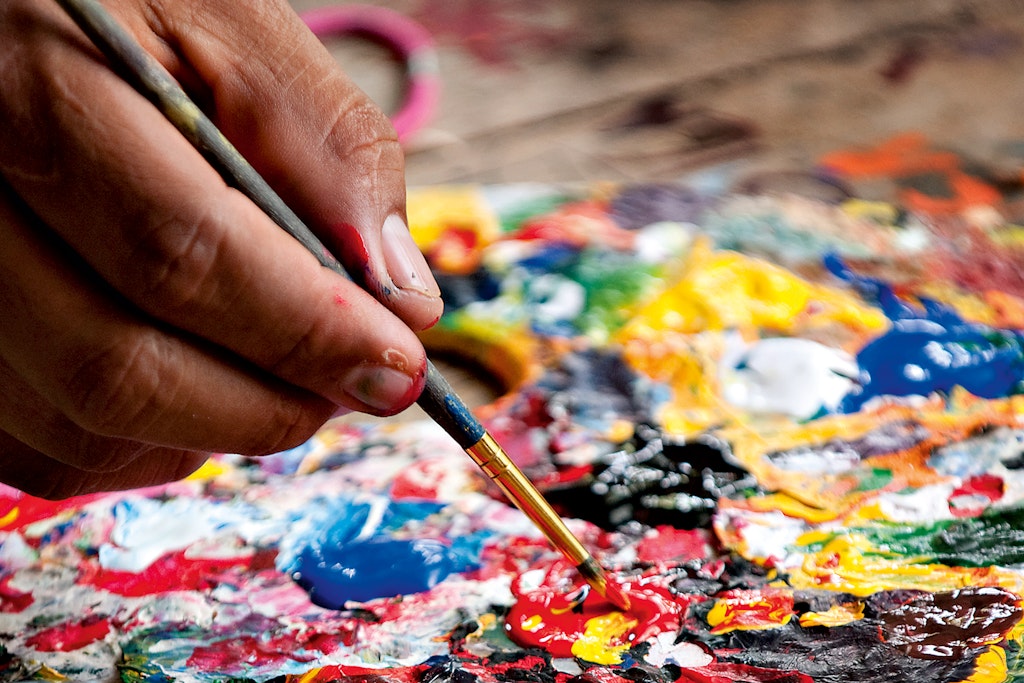 A hand holds a brush, dipping it into fresh paint.