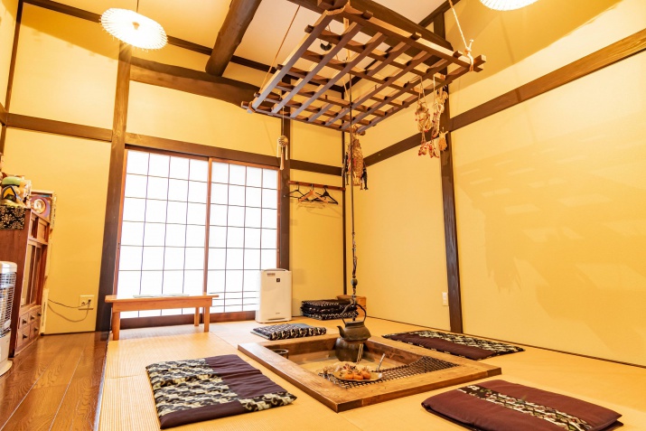A traditional tatami room with a sunken hearth, floor cushions, and a wooden hanging rack over a low table