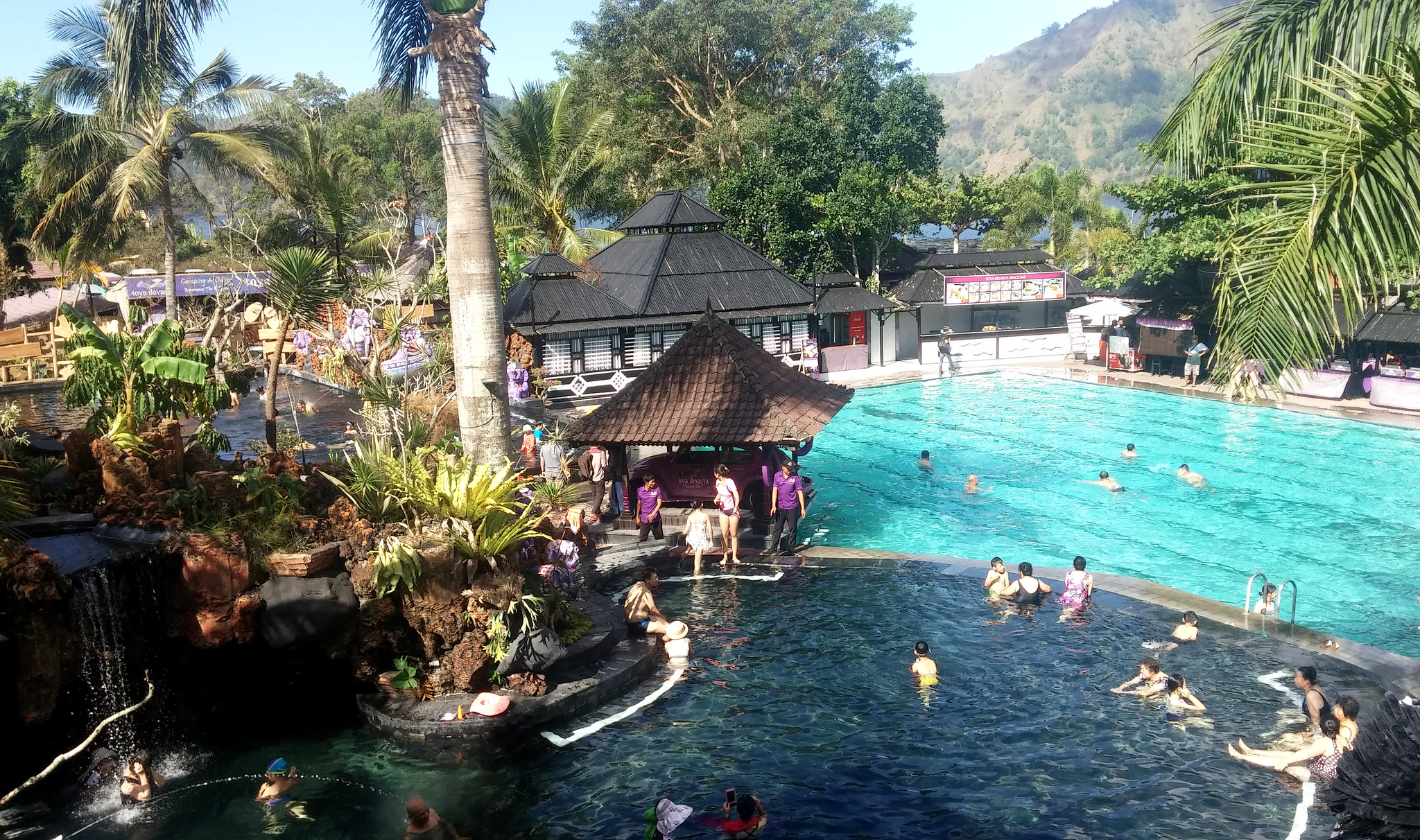 Swimmers relax in a large pool surrounded by palm trees and mountains at a Bali hot springs resort