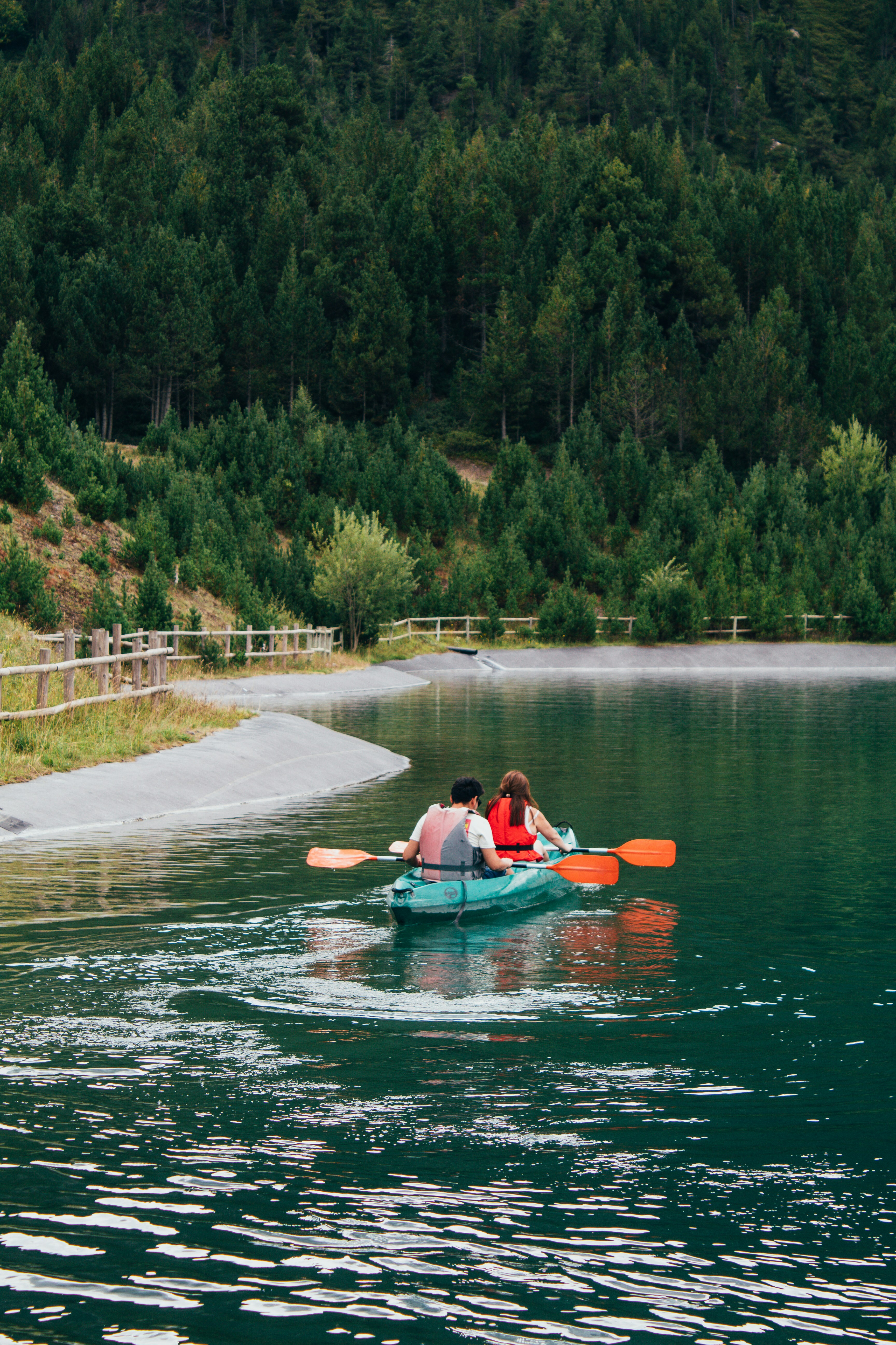 Two people paddle a green kayak across a still lake below a steep pine-covered slope.