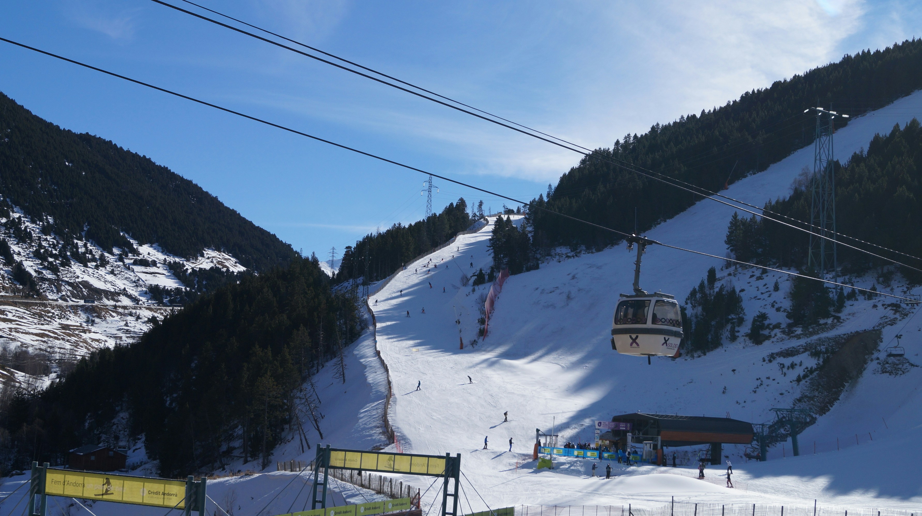 A gondola cabin climbs above a groomed ski piste with skiers on it, between forested slopes.