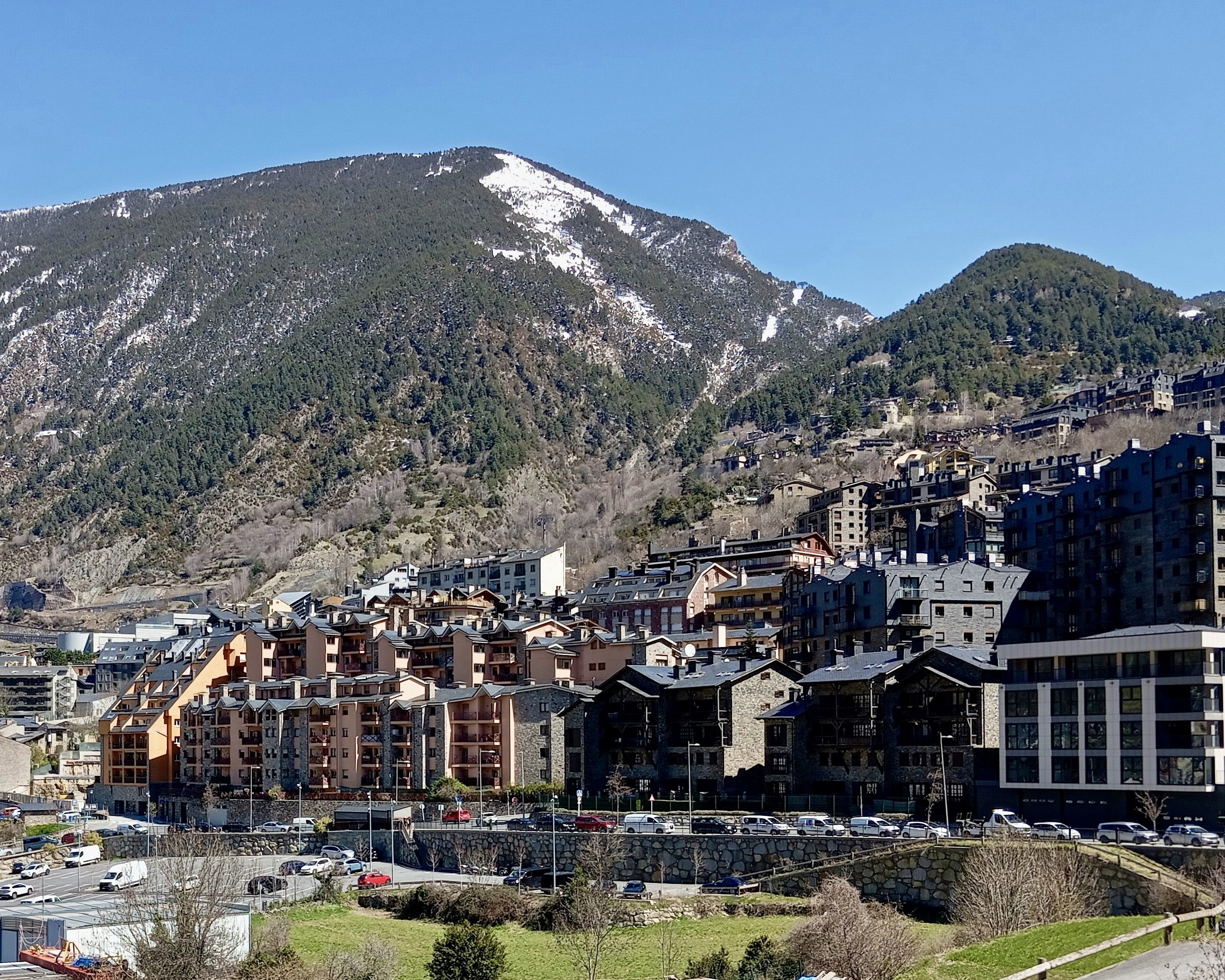 Stone and slate apartment blocks line a valley road below a pine-covered mountain with late snow on its upper slopes.