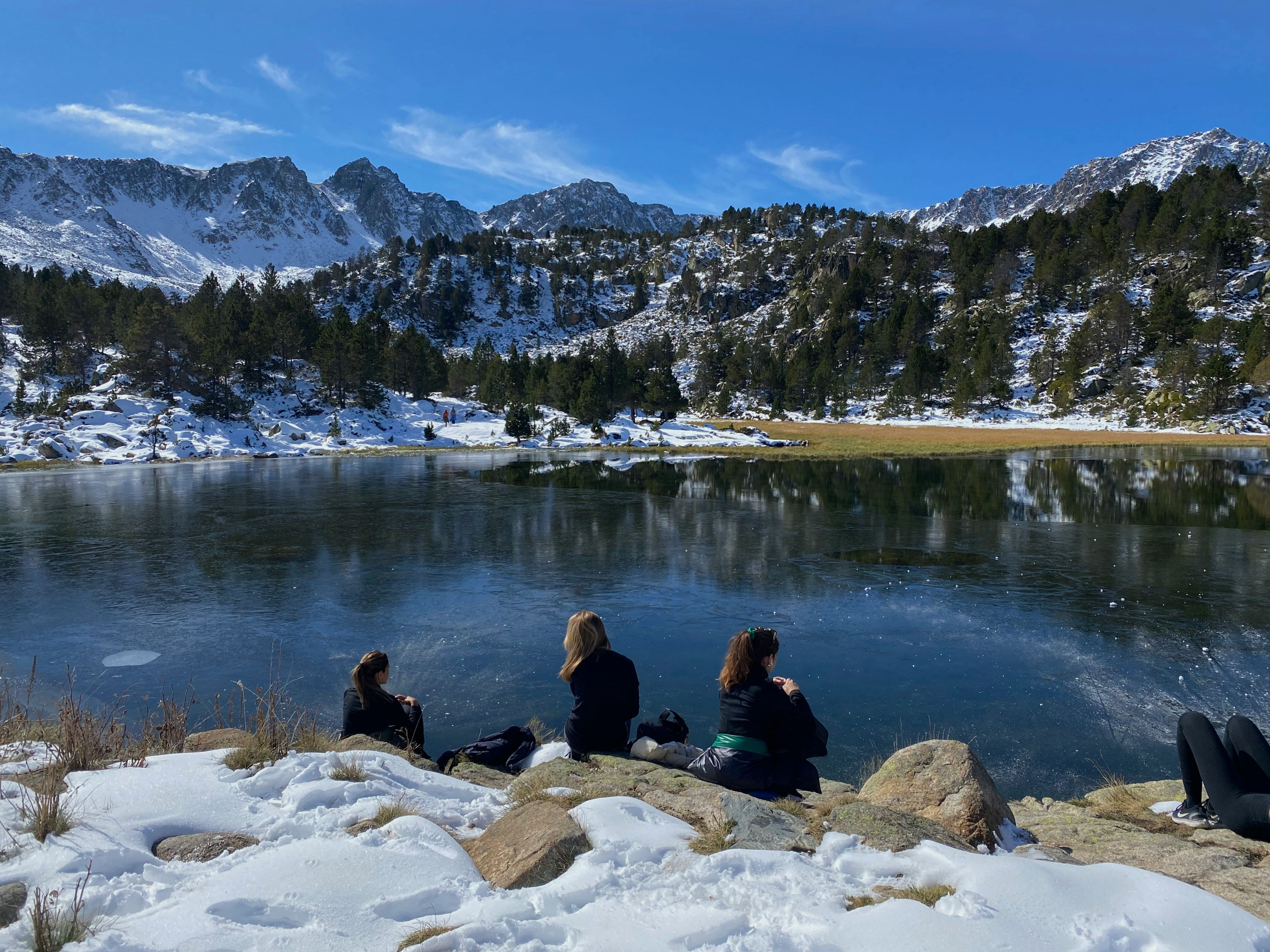 Three people sit on rocks at the edge of a frozen lake in Andorra, with snow-covered Pyrenean peaks behind them.