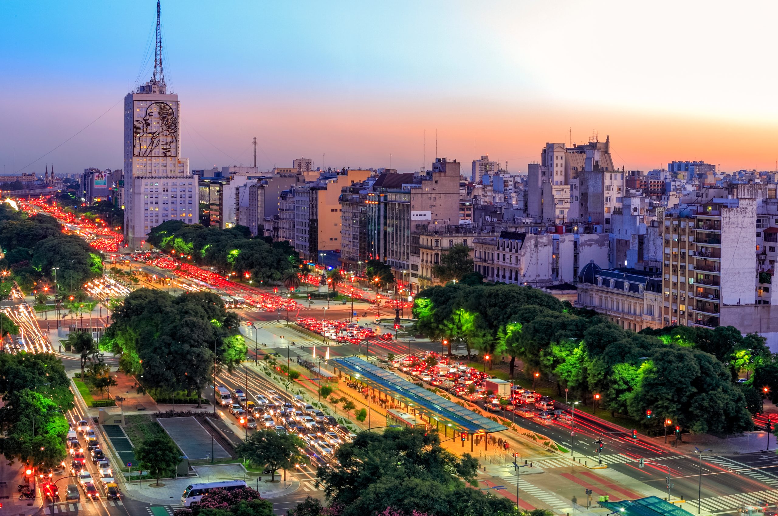 Avenida 9 de Julio in Buenos Aires at dusk, the Evita mural on the Ministry of Health building above lanes of traffic and jacaranda trees.