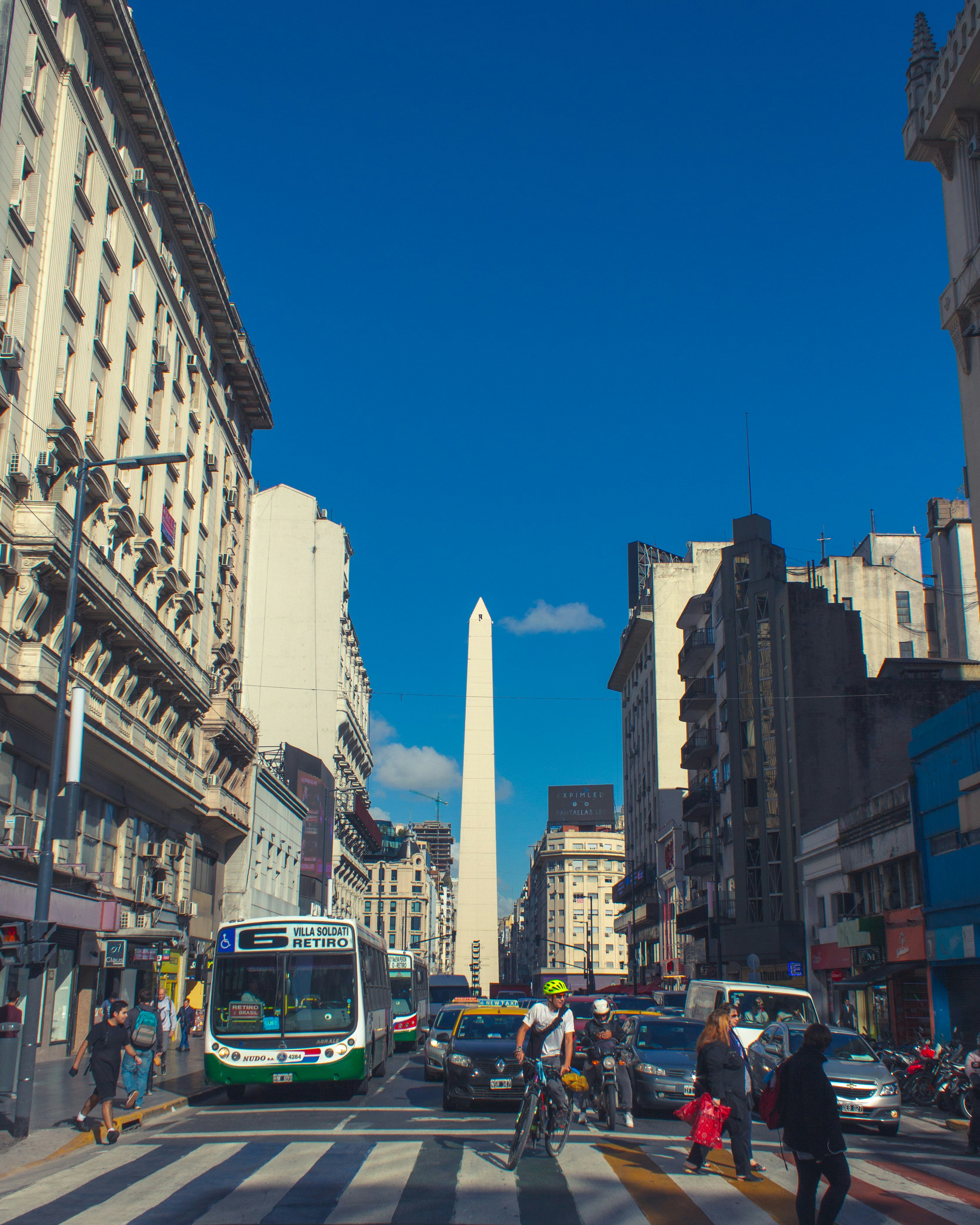 The Obelisco at the end of a busy city street, with a green city bus, taxis and pedestrians on the crossing.