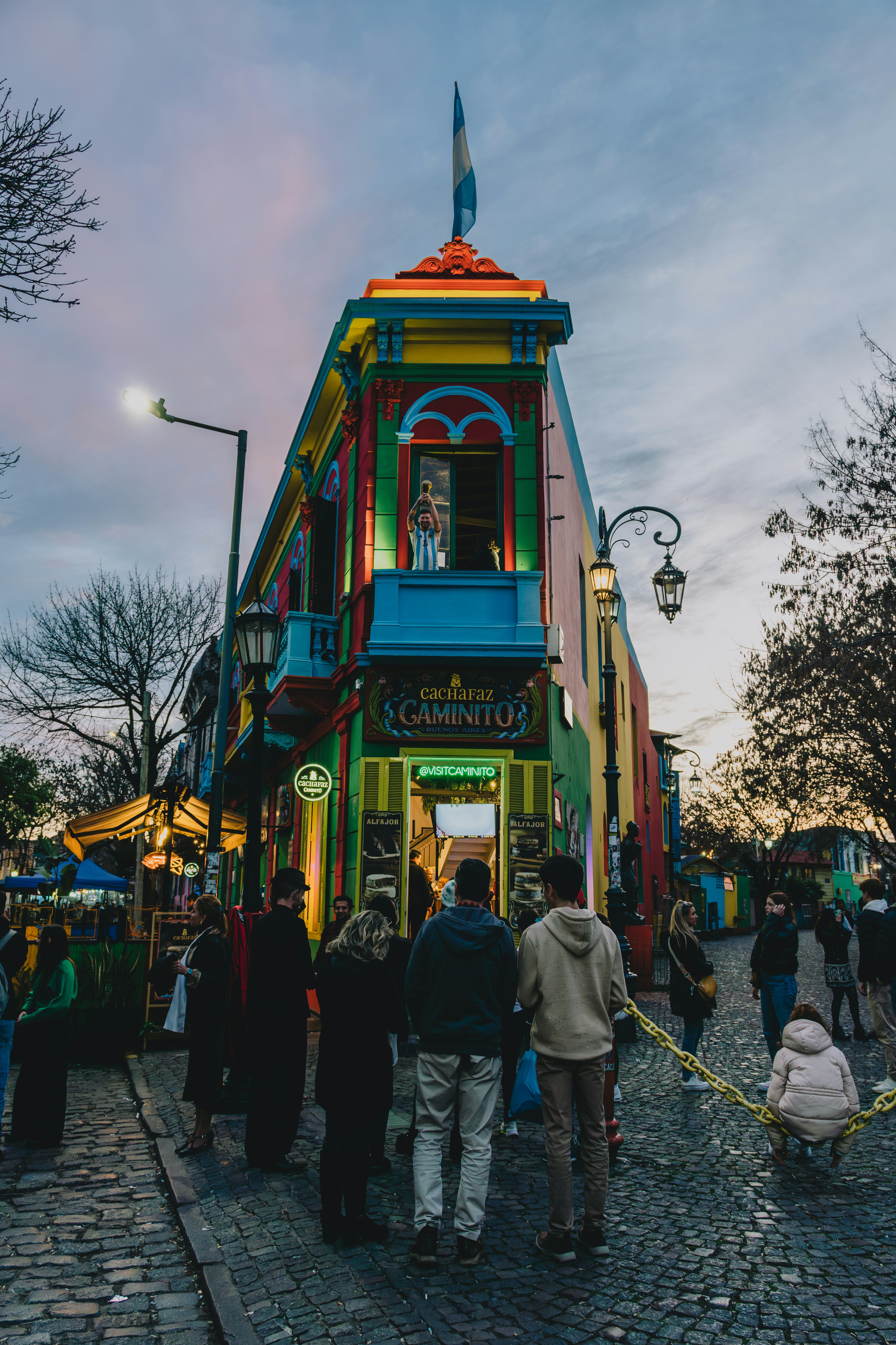 The painted corner building of Caminito in La Boca at dusk, lit from within, with a street lamp and people on the cobbles.