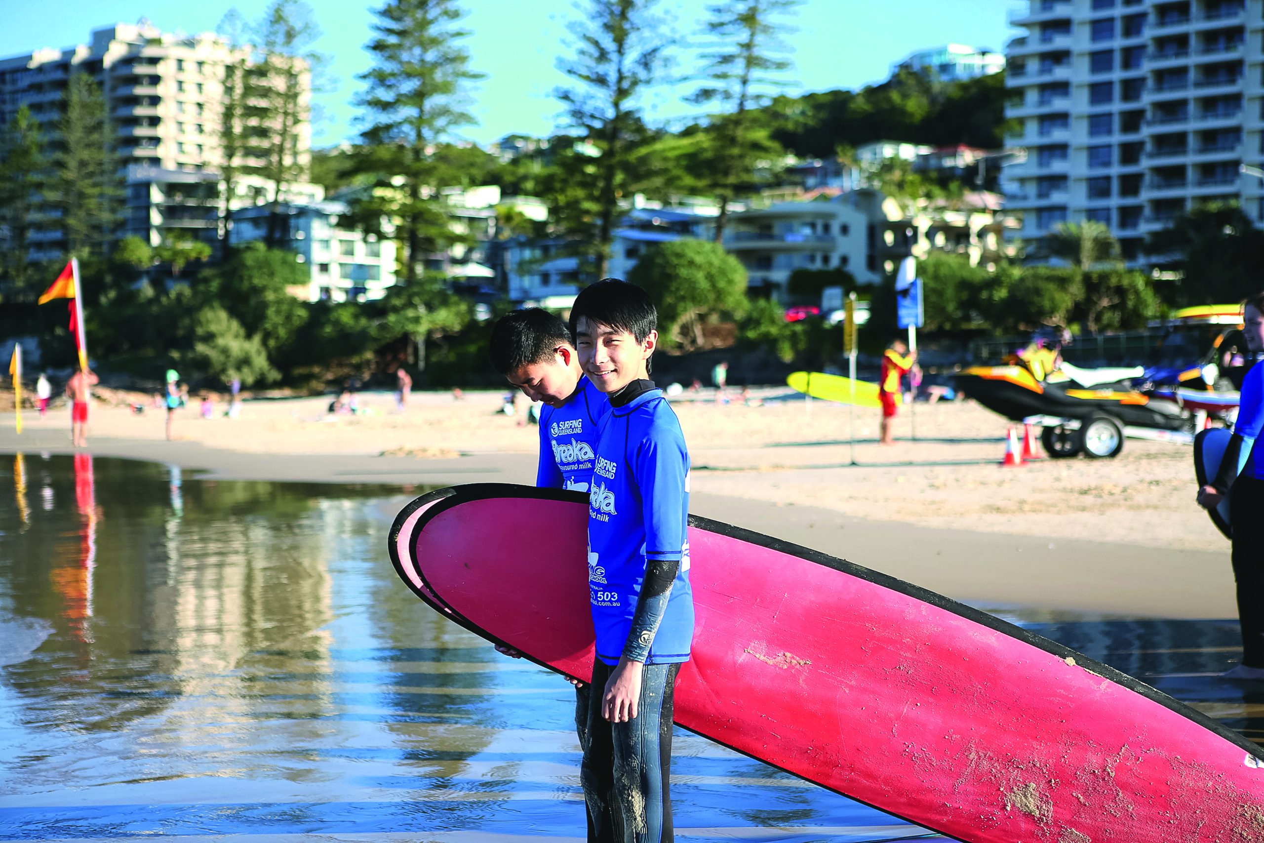 A student carries a longboard into shallow water at a surf beach, apartments behind