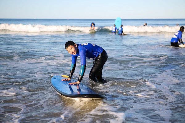 A teen traveler in a rash guard kneels on a soft-top surfboard in the shallows as small waves roll in
