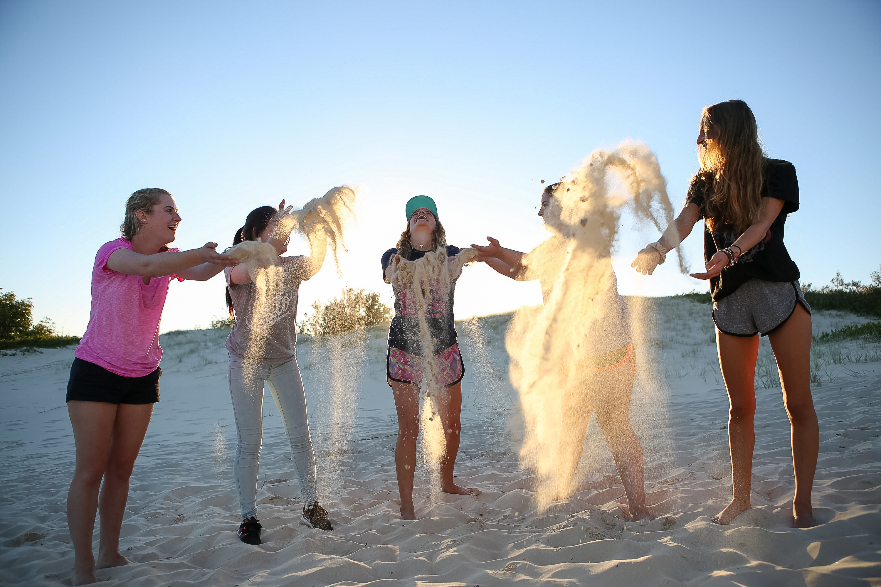 Students throw sand into the air while playing on a beach at sunset