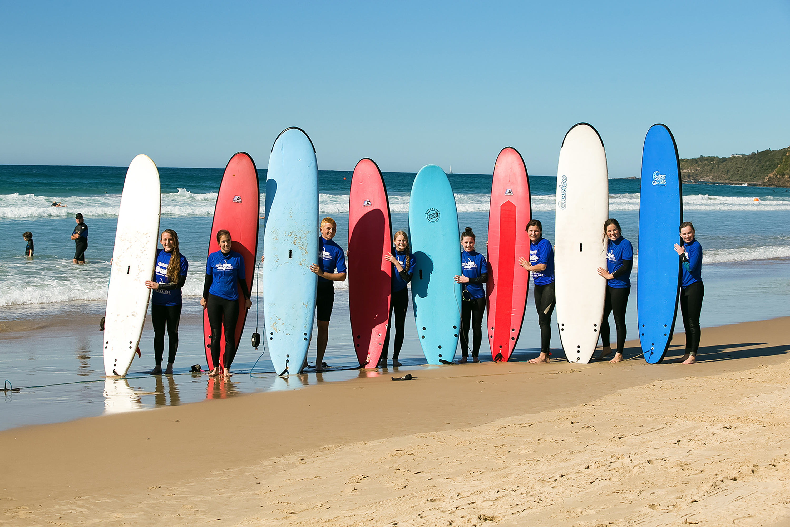 Students in wetsuits line up on the beach holding surfboards upright
