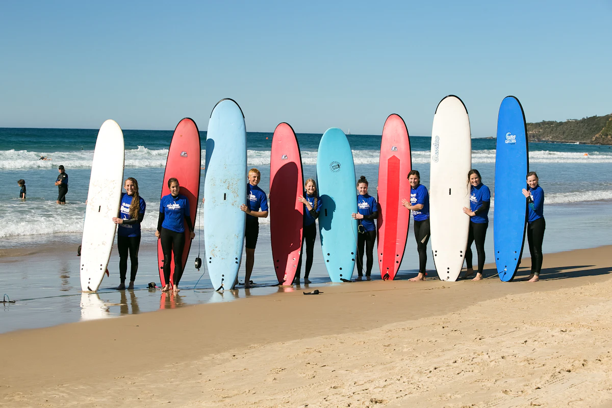 Teen travelers in blue rash guards stand between a row of upright surfboards on a sunny beach in Australia