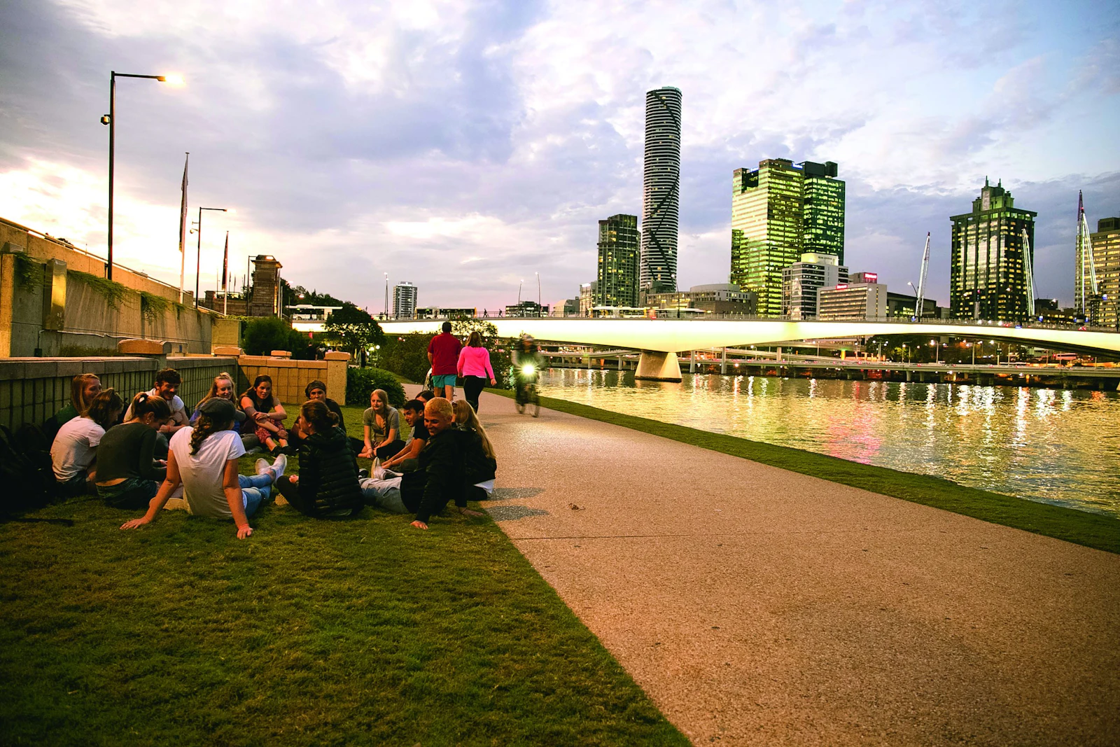 Teen travelers sit on the grass beside a riverside path, with the Brisbane skyline and a bridge across the river behind them