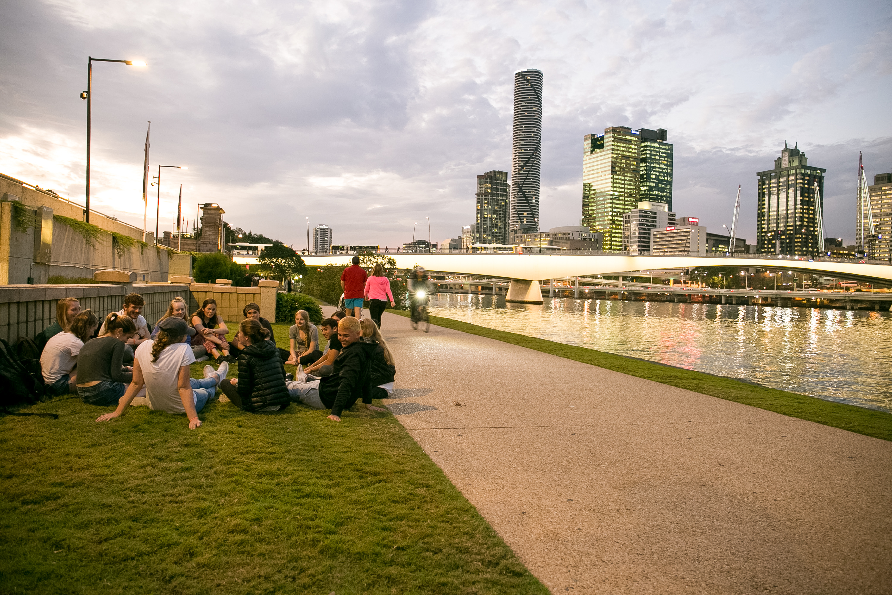 Students in Australia sit on riverside grass with a city skyline and bridge across the water at dusk