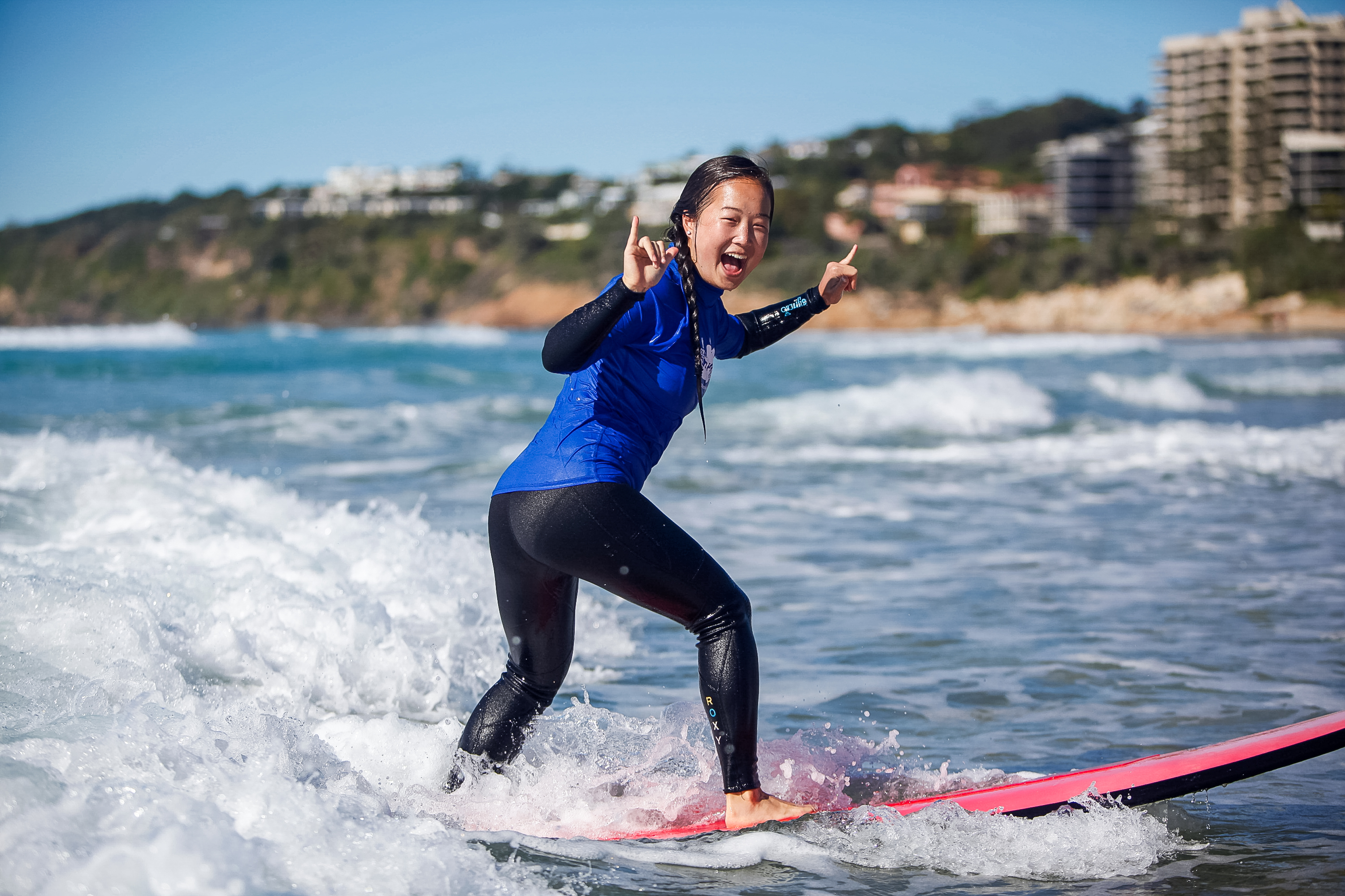 A student balances on a surfboard while riding a wave toward shore, coastal buildings in the background