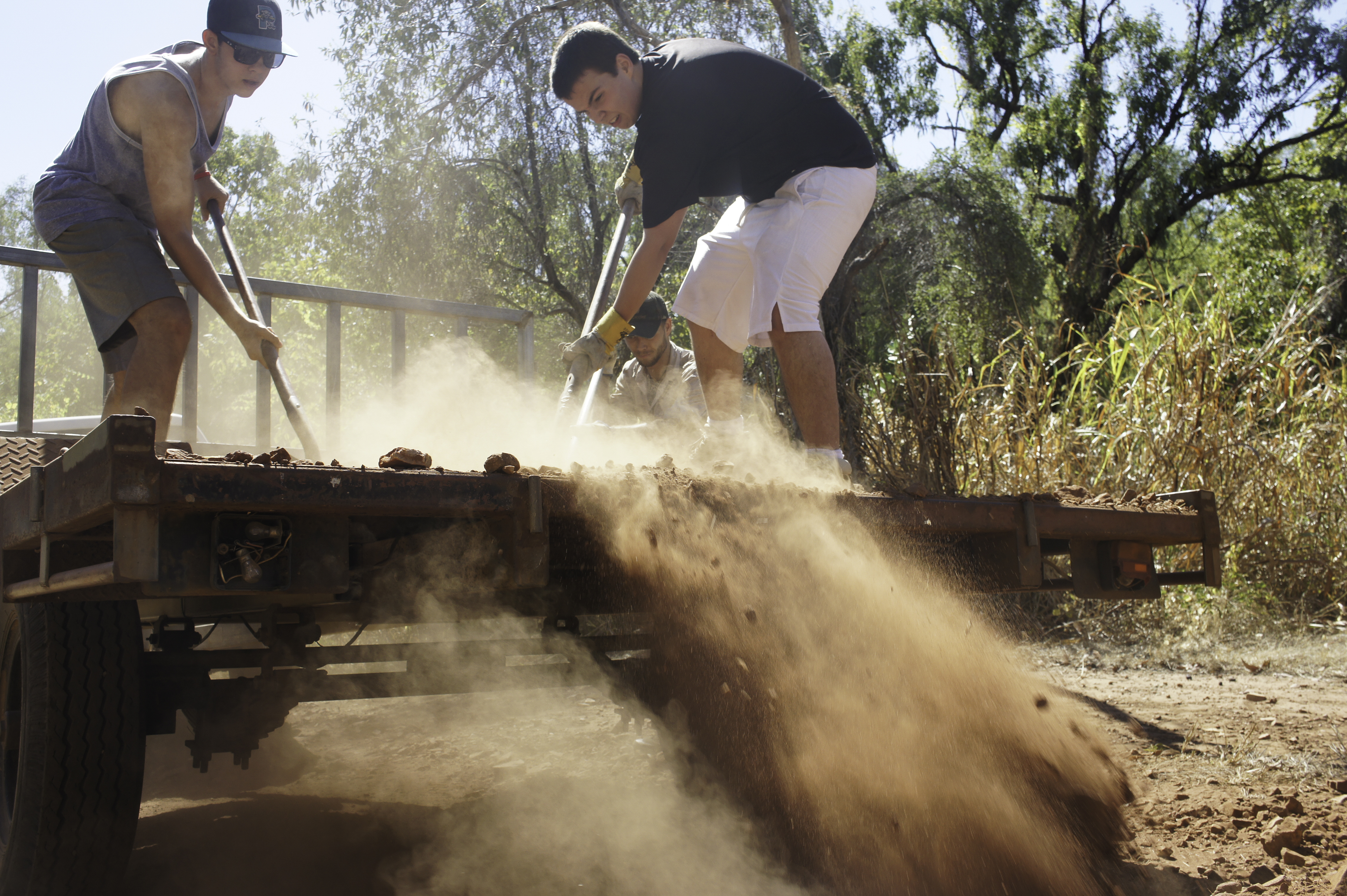 Students shovel dirt off a flatbed trailer in a cloud of dust