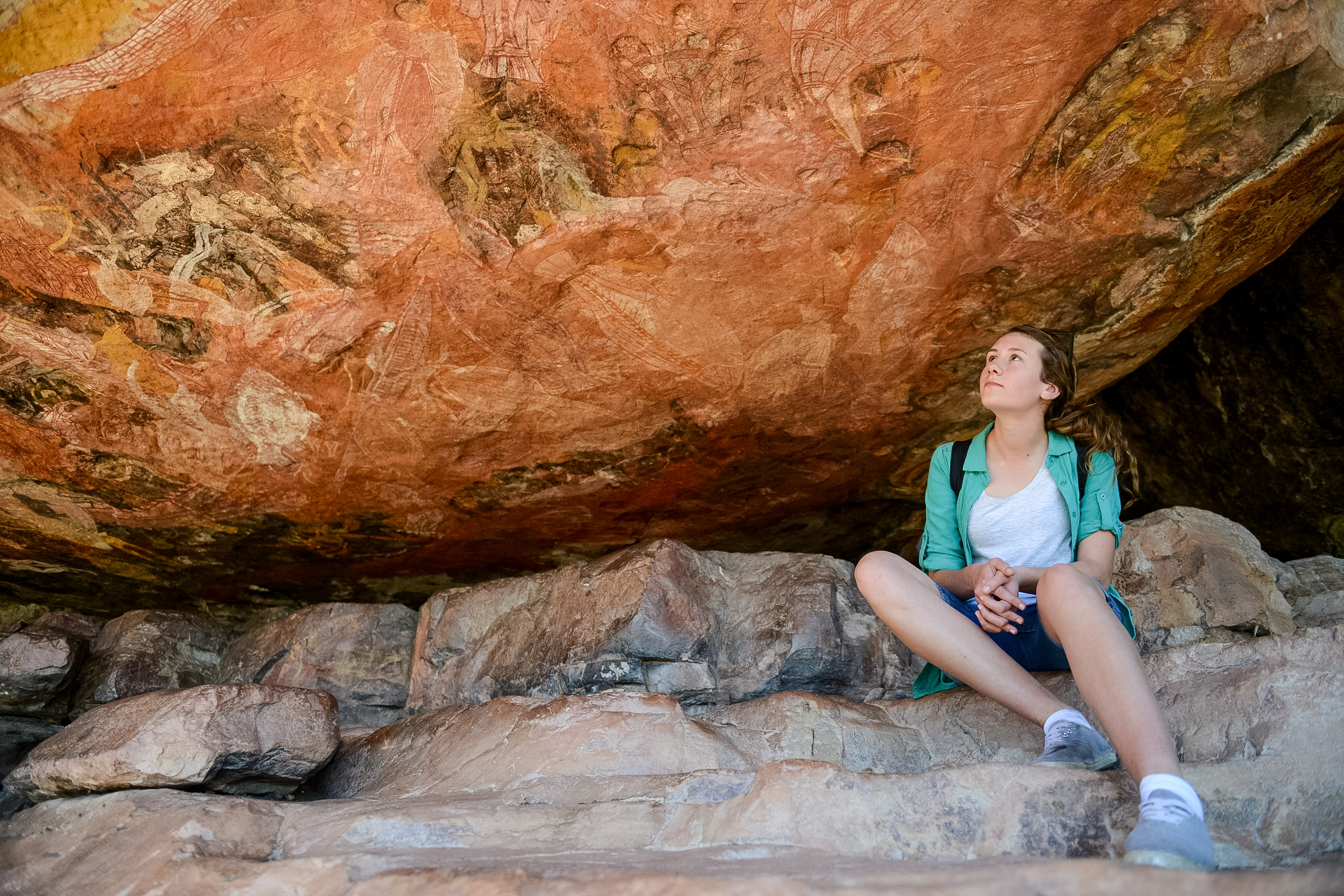 On a school trip, a student sits on rocks beneath ochre-colored Indigenous rock art in a cave overhang