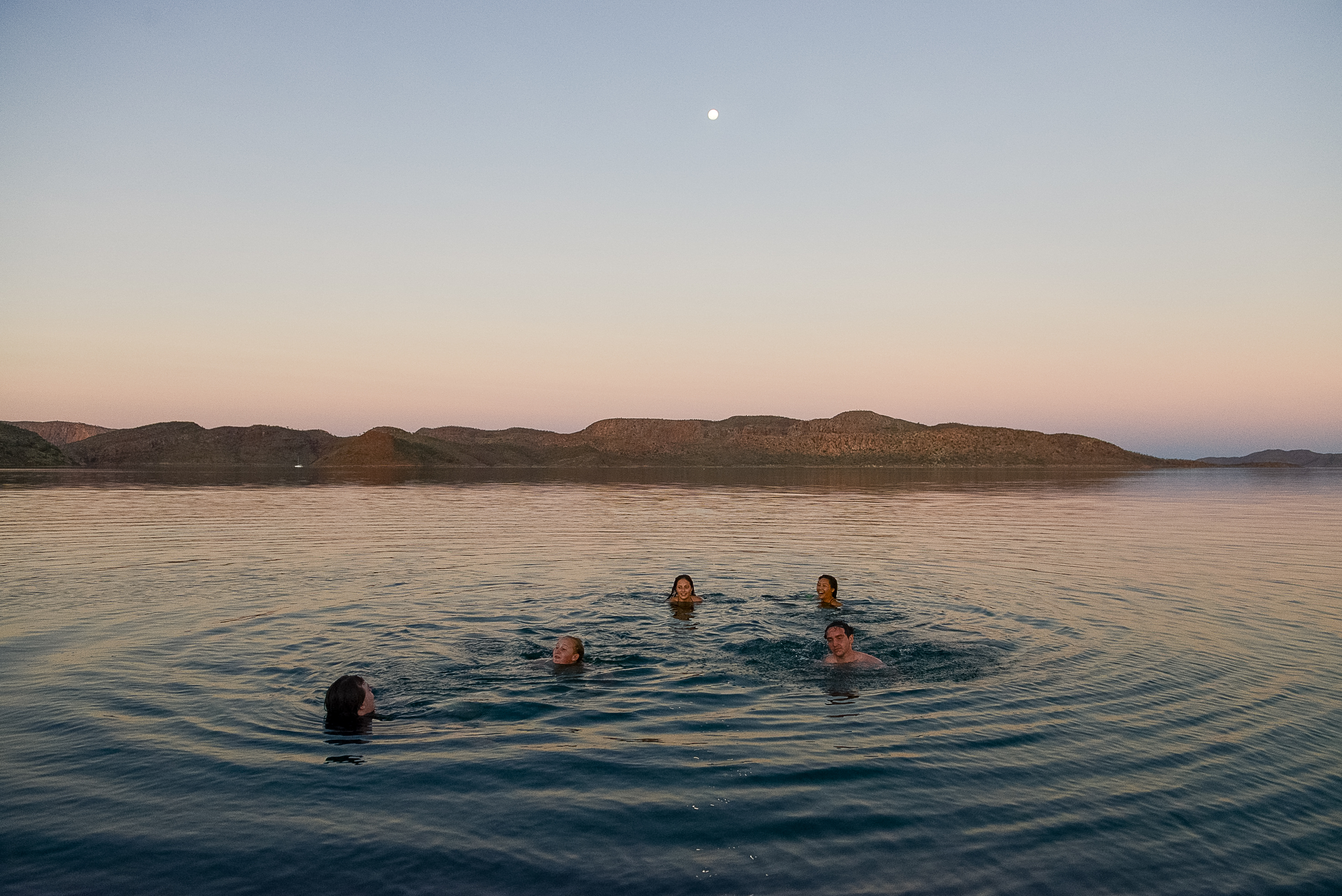 Students swim in calm water at dusk with hills in the distance and the moon overhead