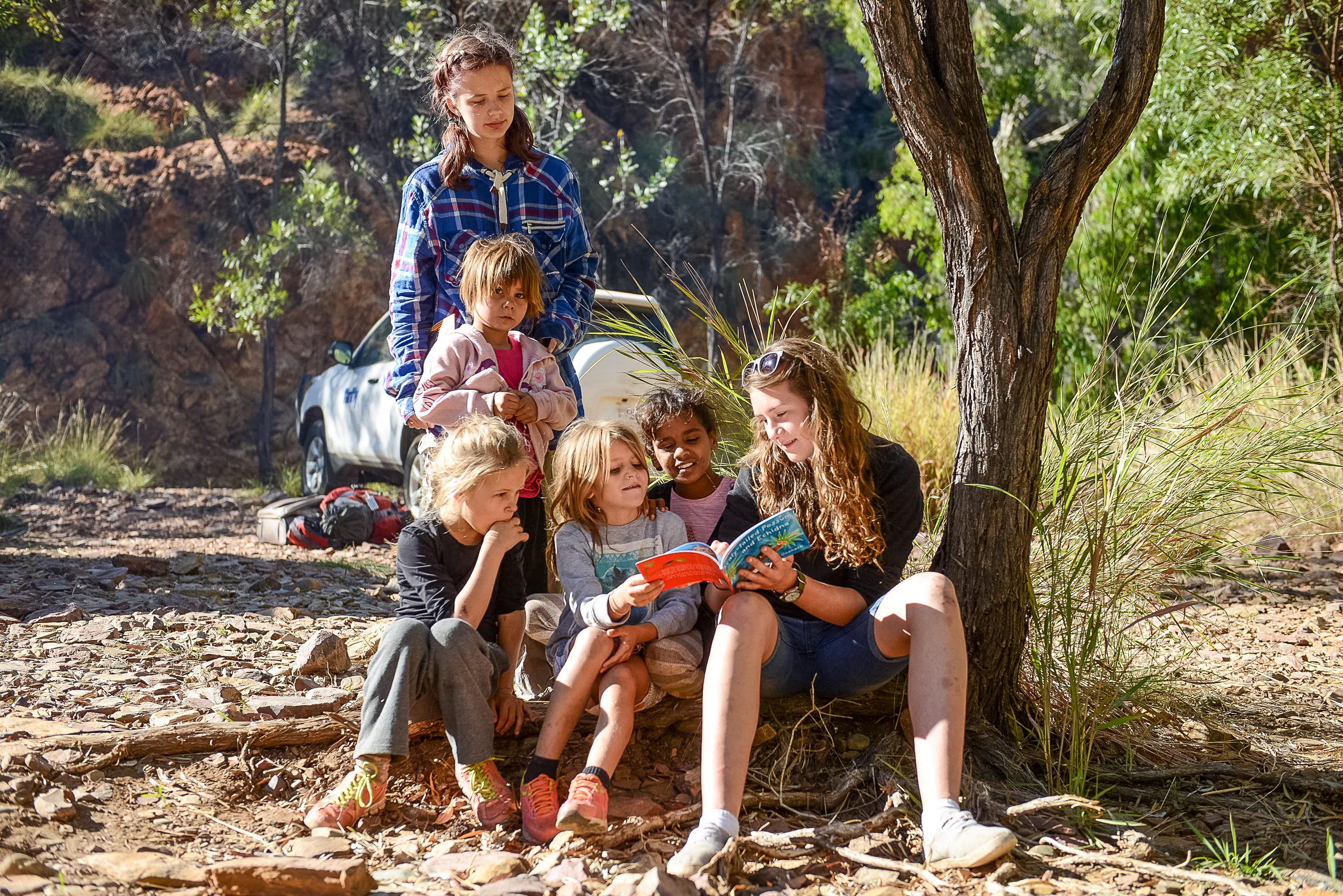 Students sit under a tree reading a picture book with young children