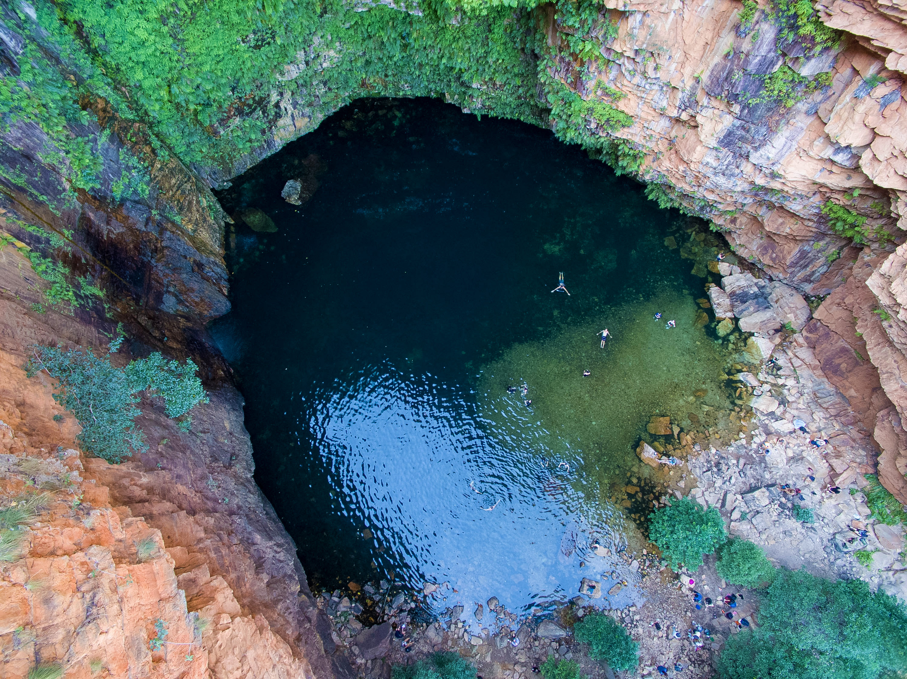 Aerial view of swimmers in a deep, rock-walled waterhole surrounded by greenery