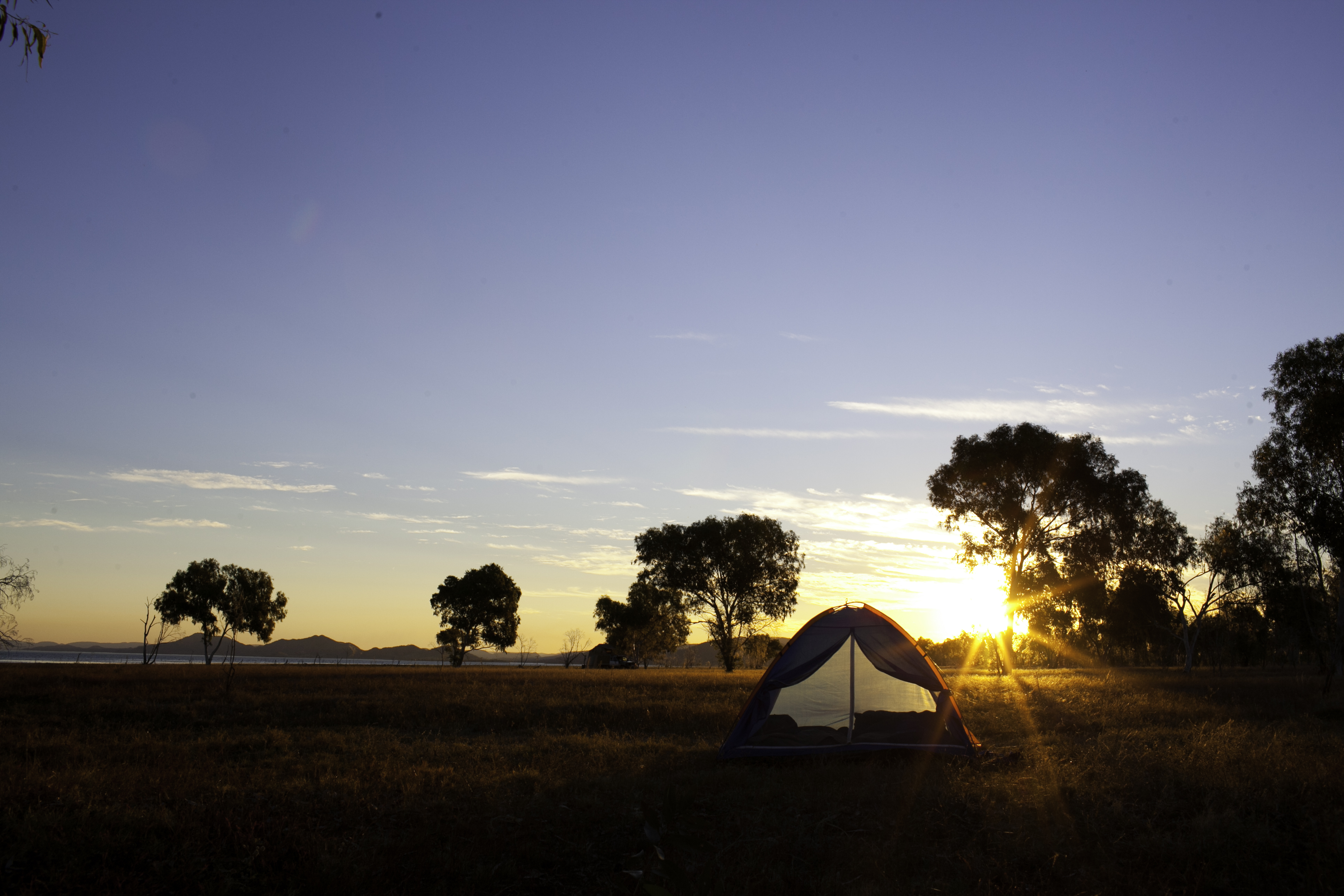 A tent glows in silhouette against the sunset with scattered trees on the horizon