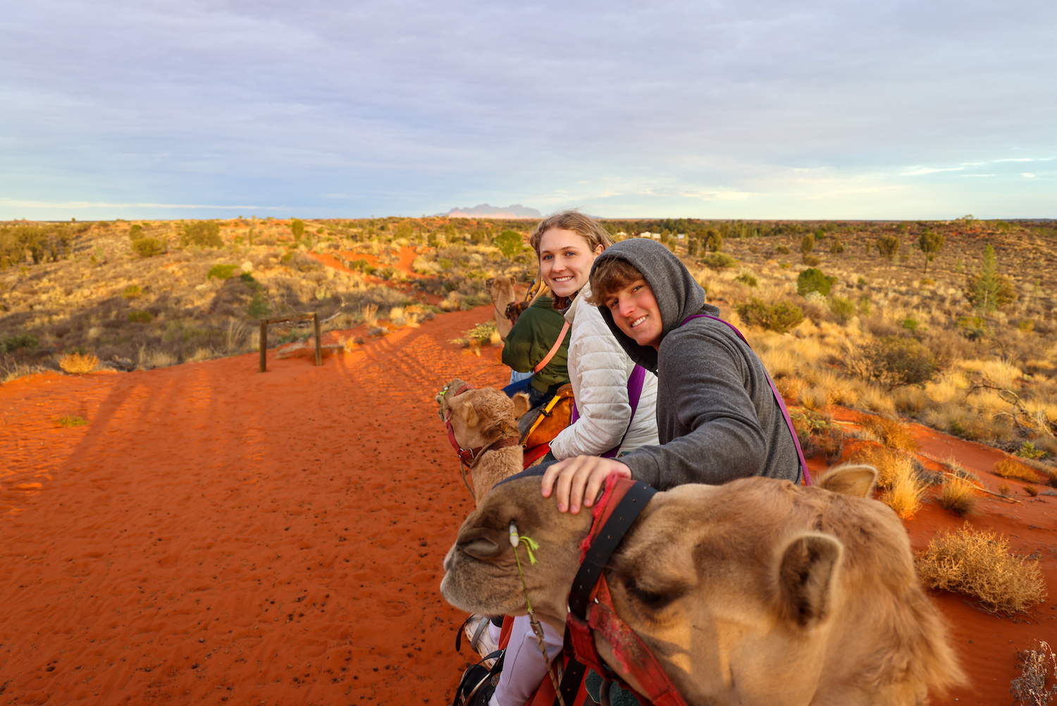 Two people riding camels in a desert at sunset.