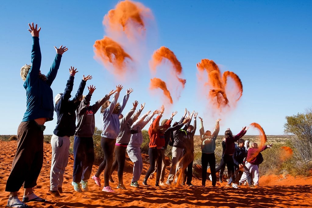 Group of people throwing red sand into the air in a desert.