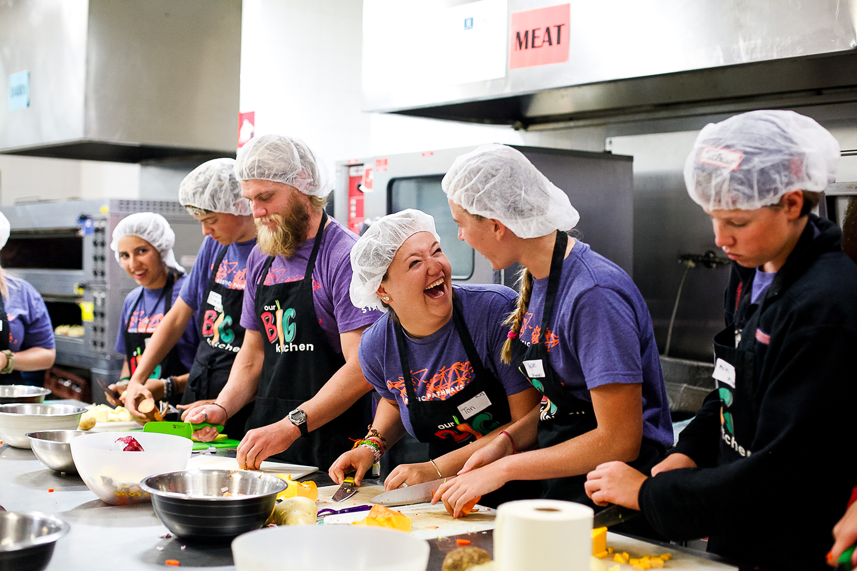 Teens in hairnets laugh while chopping vegetables in a commercial kitchen