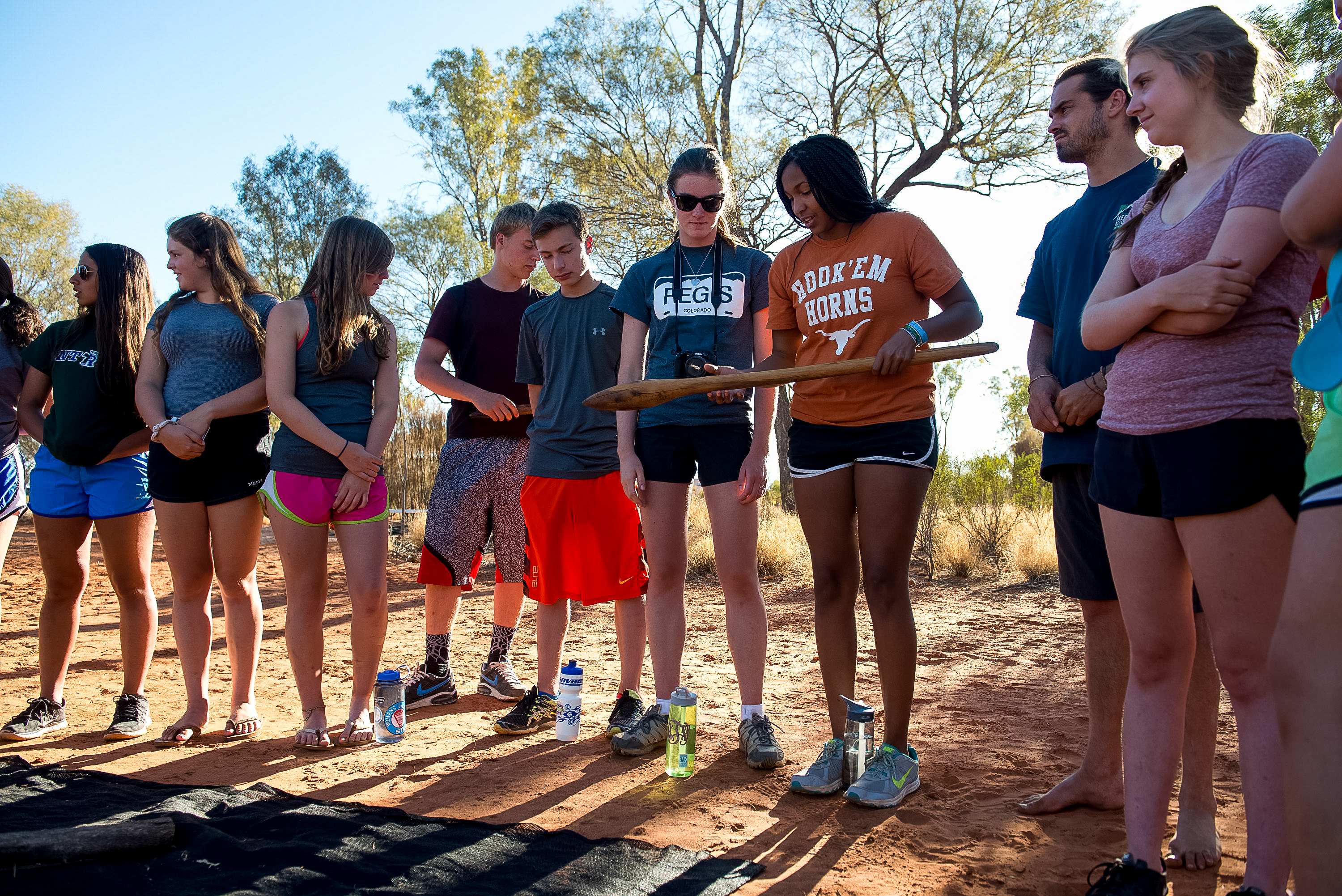 Students stand in a line outdoors holding a wooden stick during an activity in the outback