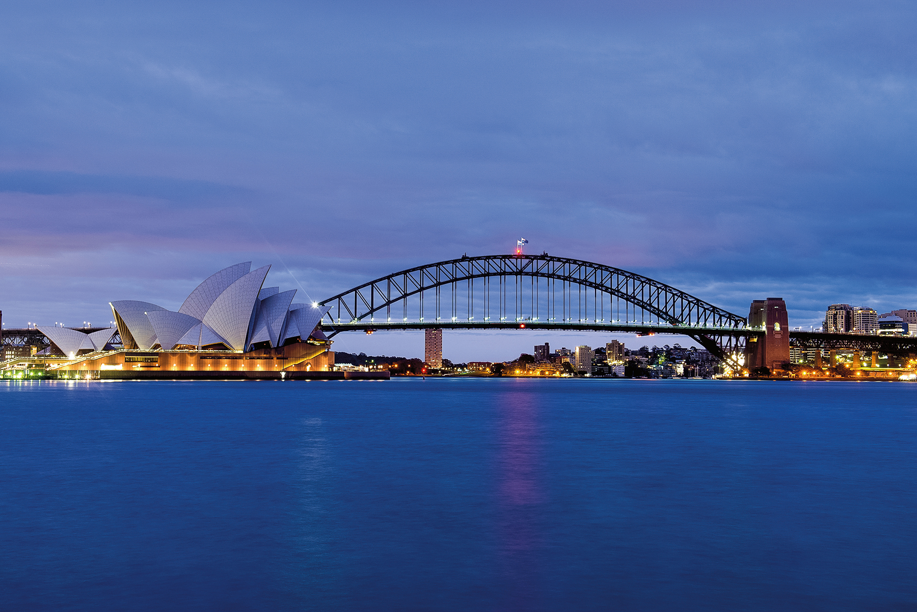 Students sit on the grass by a river with a city skyline across the water