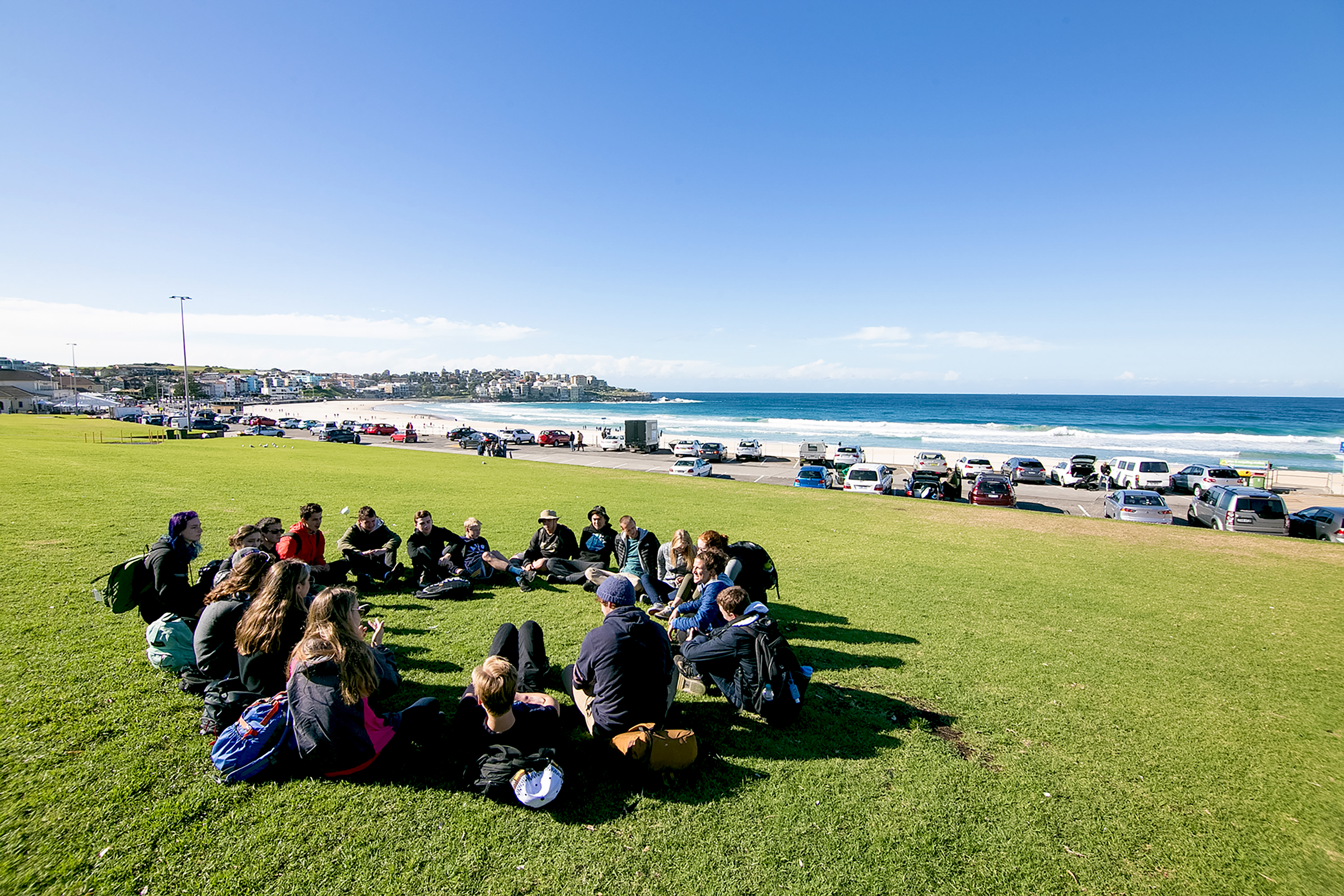 Students sit in a circle on a coastal lawn overlooking the beach and parked cars