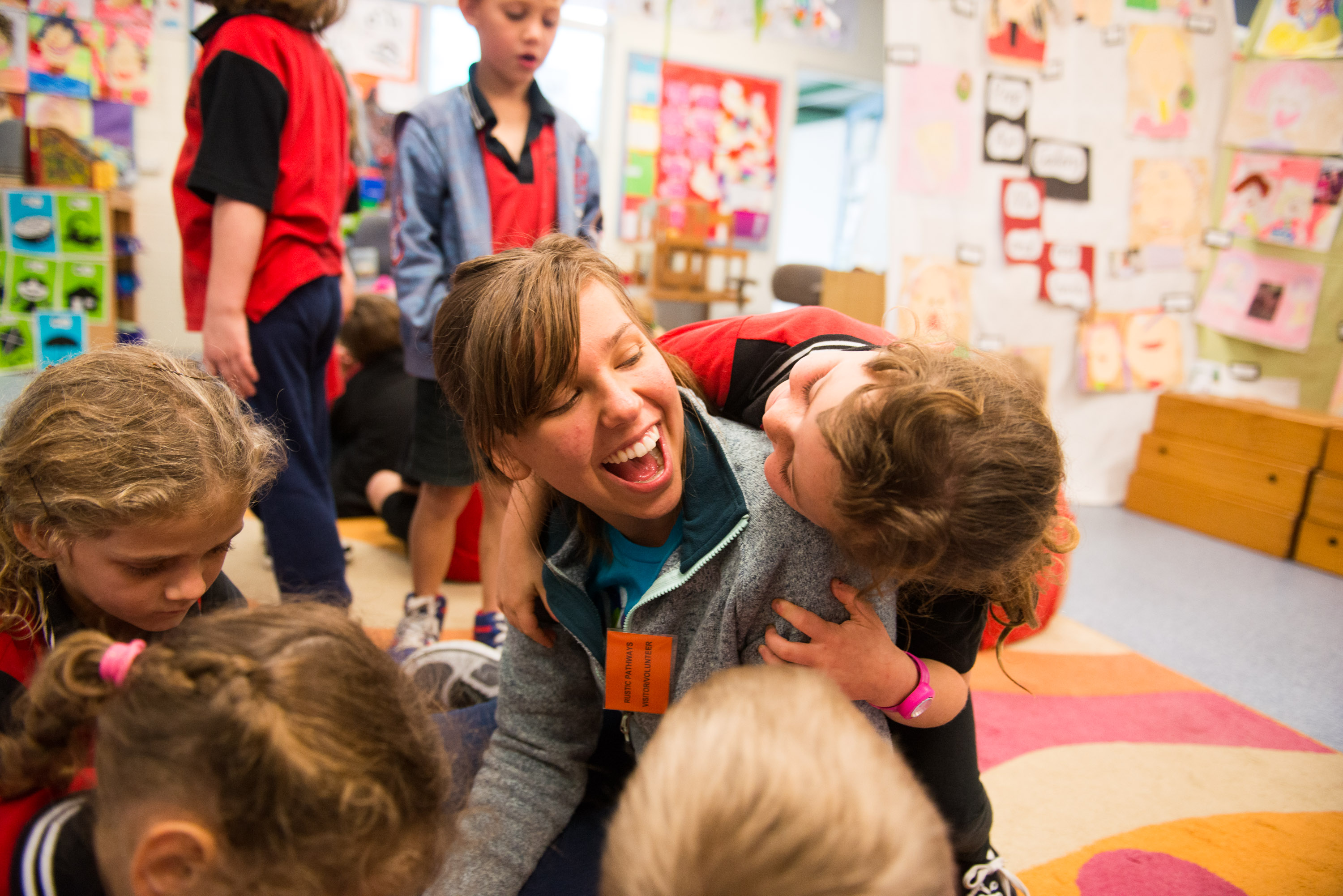 Students laugh as young children hug and climb on them in a classroom