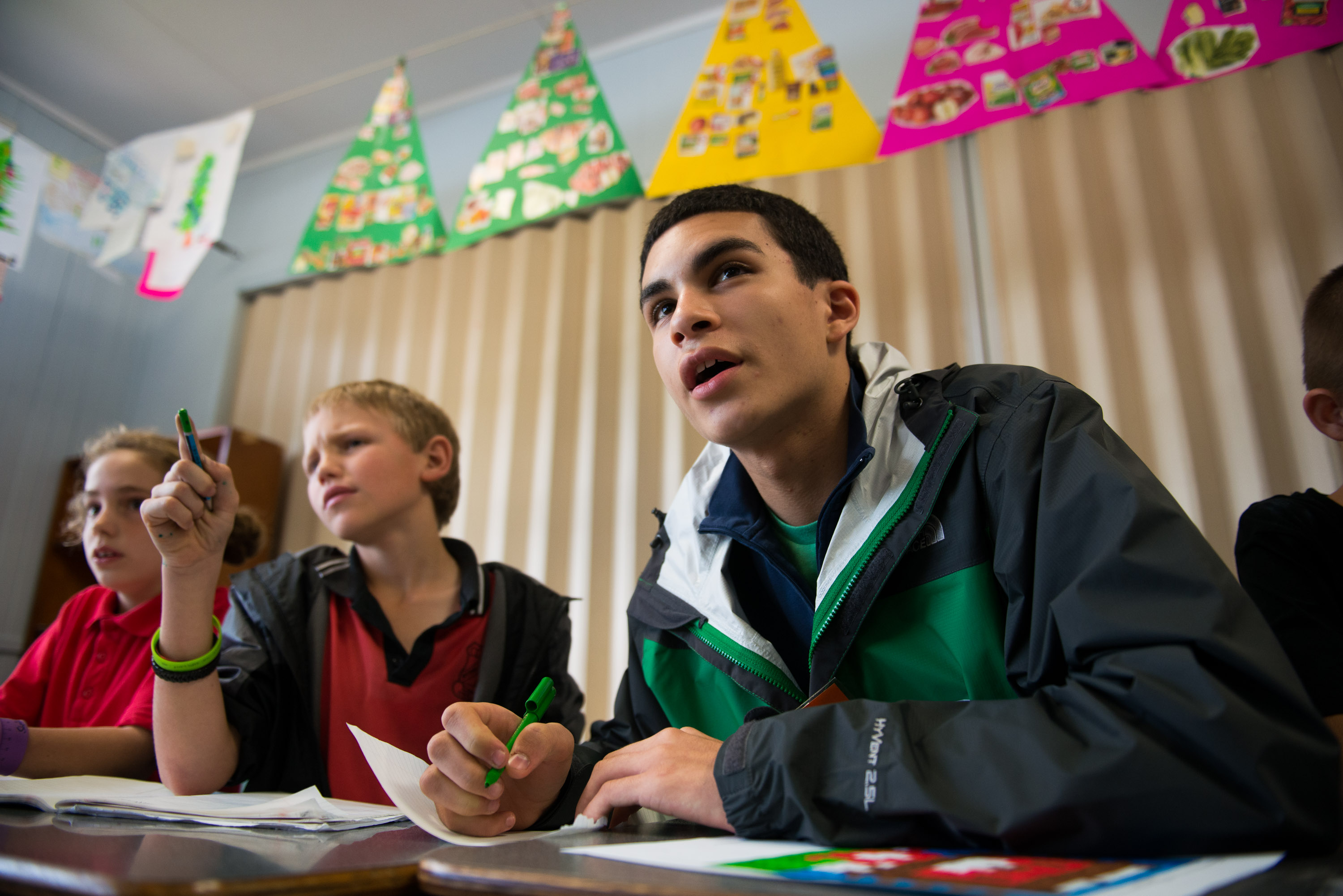 A student speaks up at a classroom desk beside primary school children
