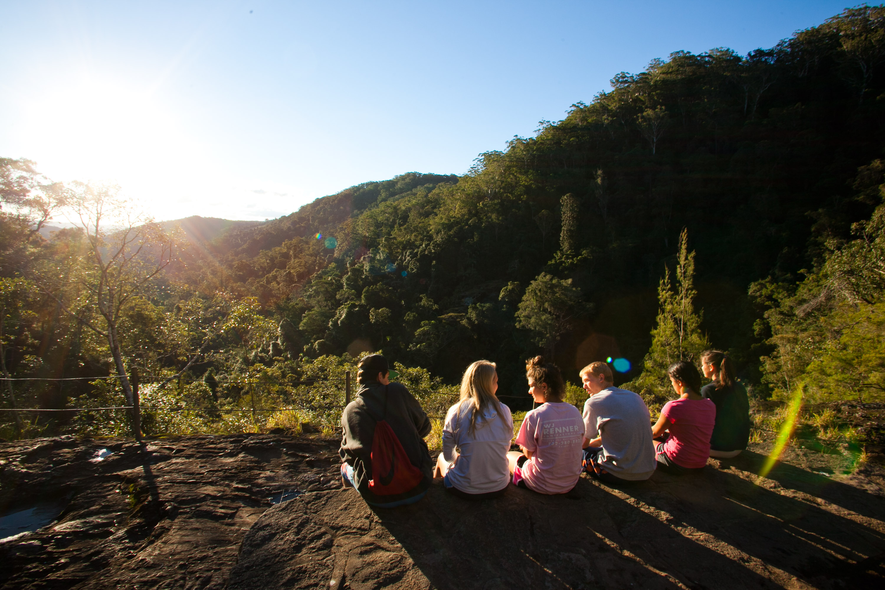 Students sit along a rocky ledge overlooking a forested valley at sunset