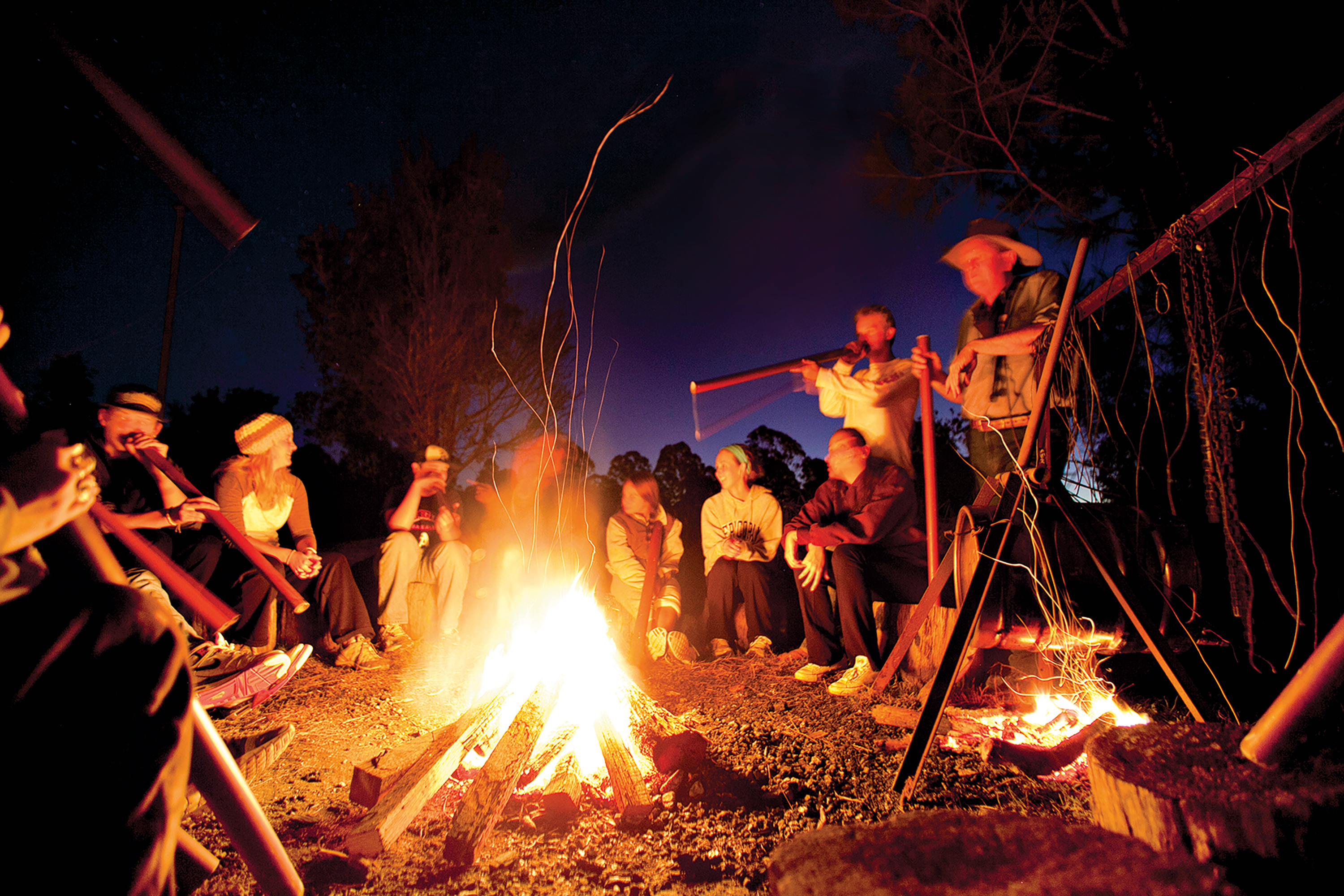 Students gather around a campfire at night while a performer plays a didgeridoo
