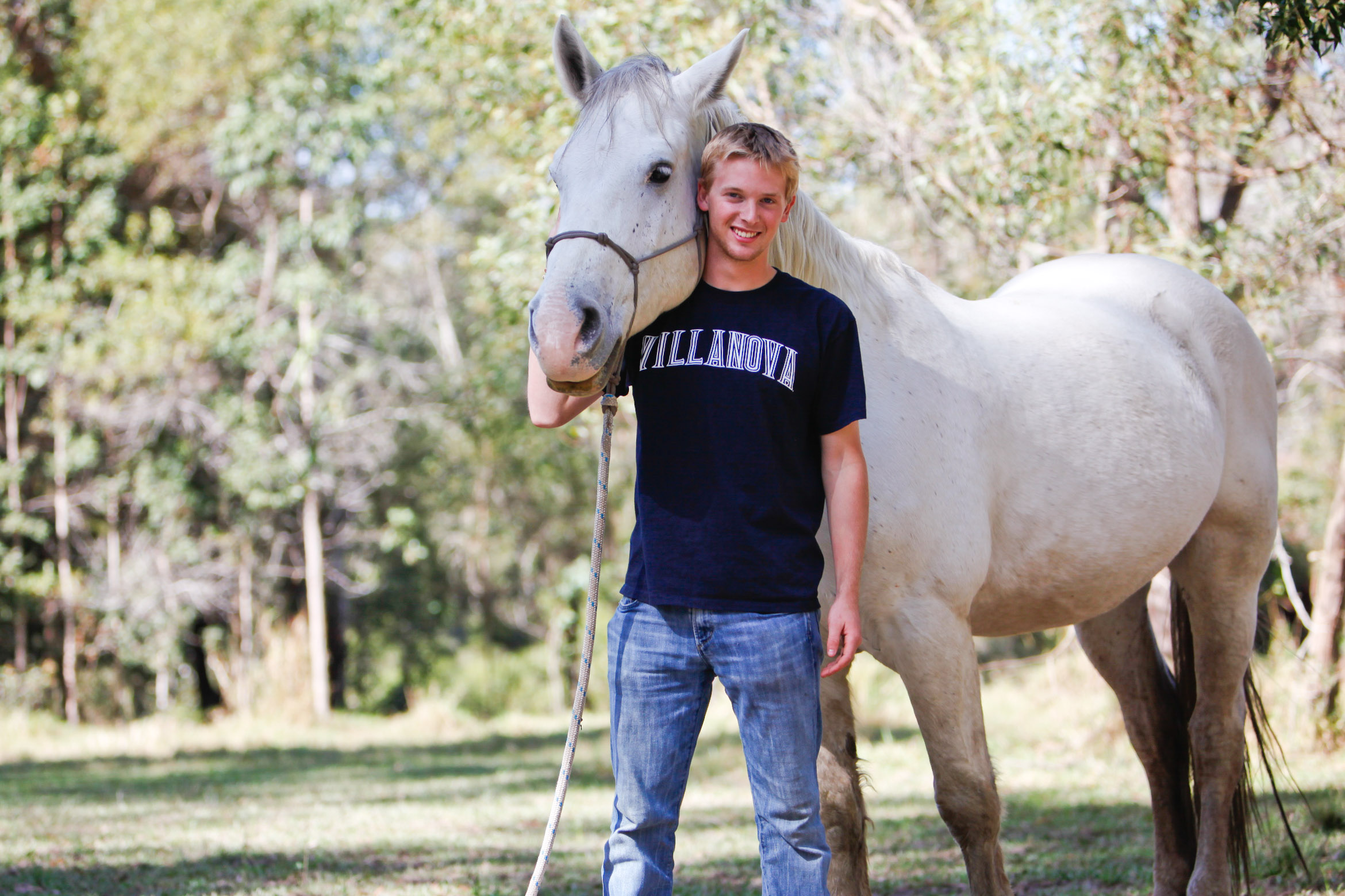 A student stands beside a white horse in a grassy, tree-lined field