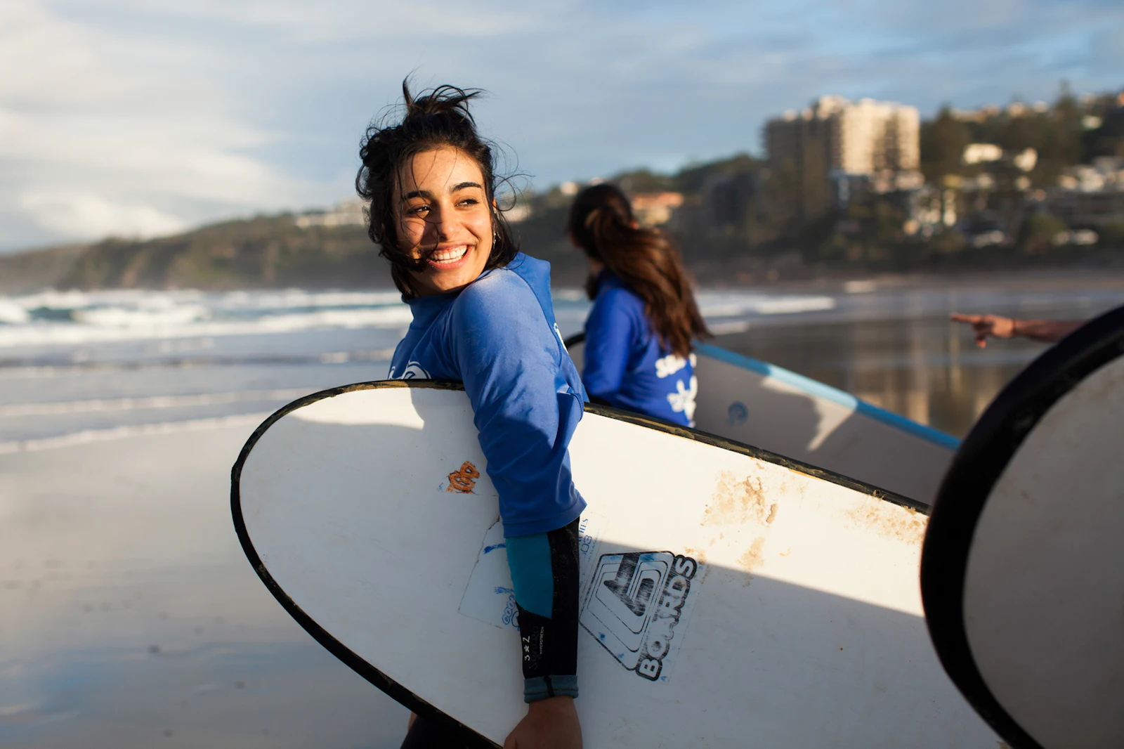 A teen traveler smiles over her shoulder as she carries a surfboard along a beach