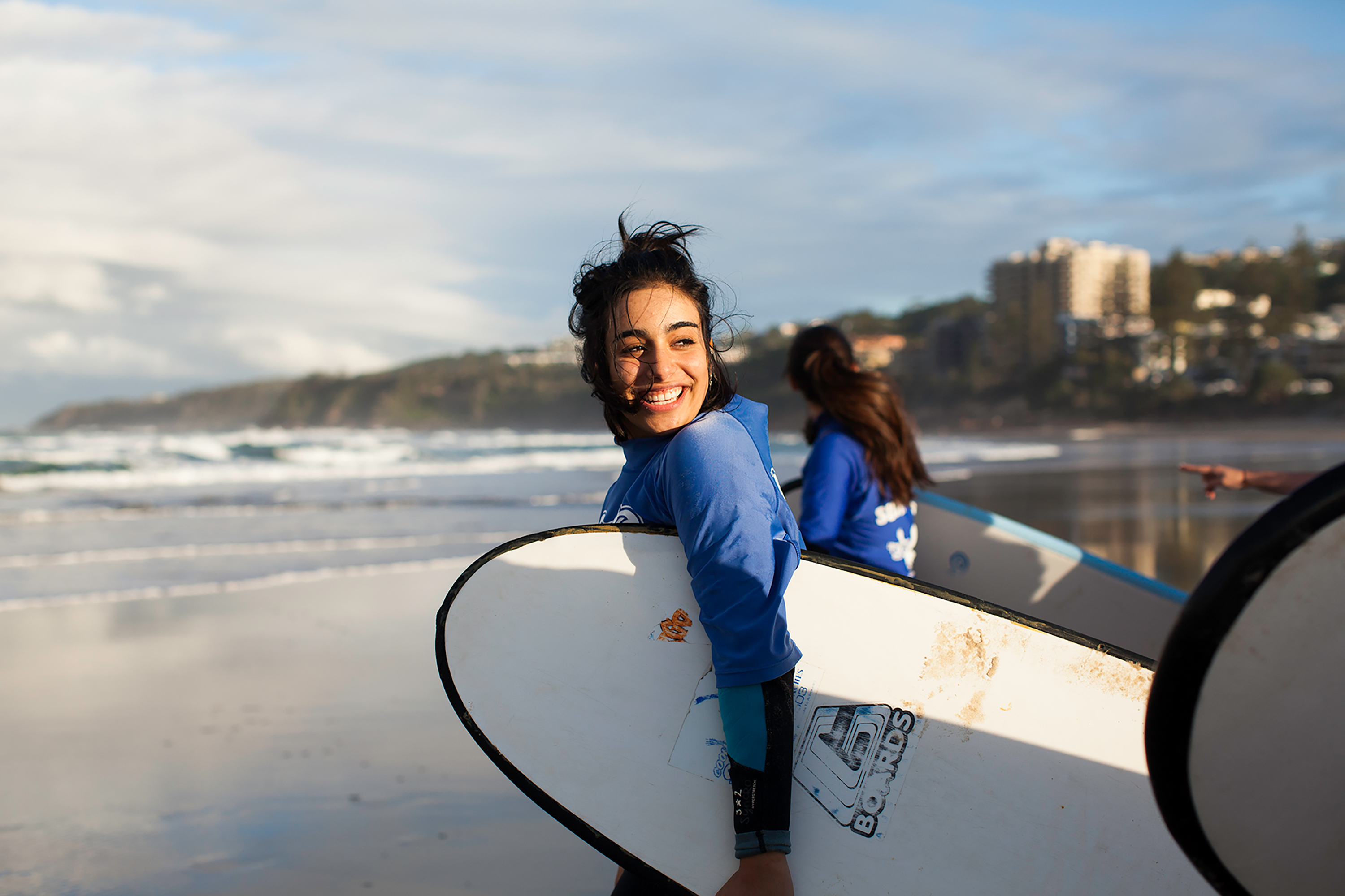 A student in Australia smiles while carrying a surfboard along the beach