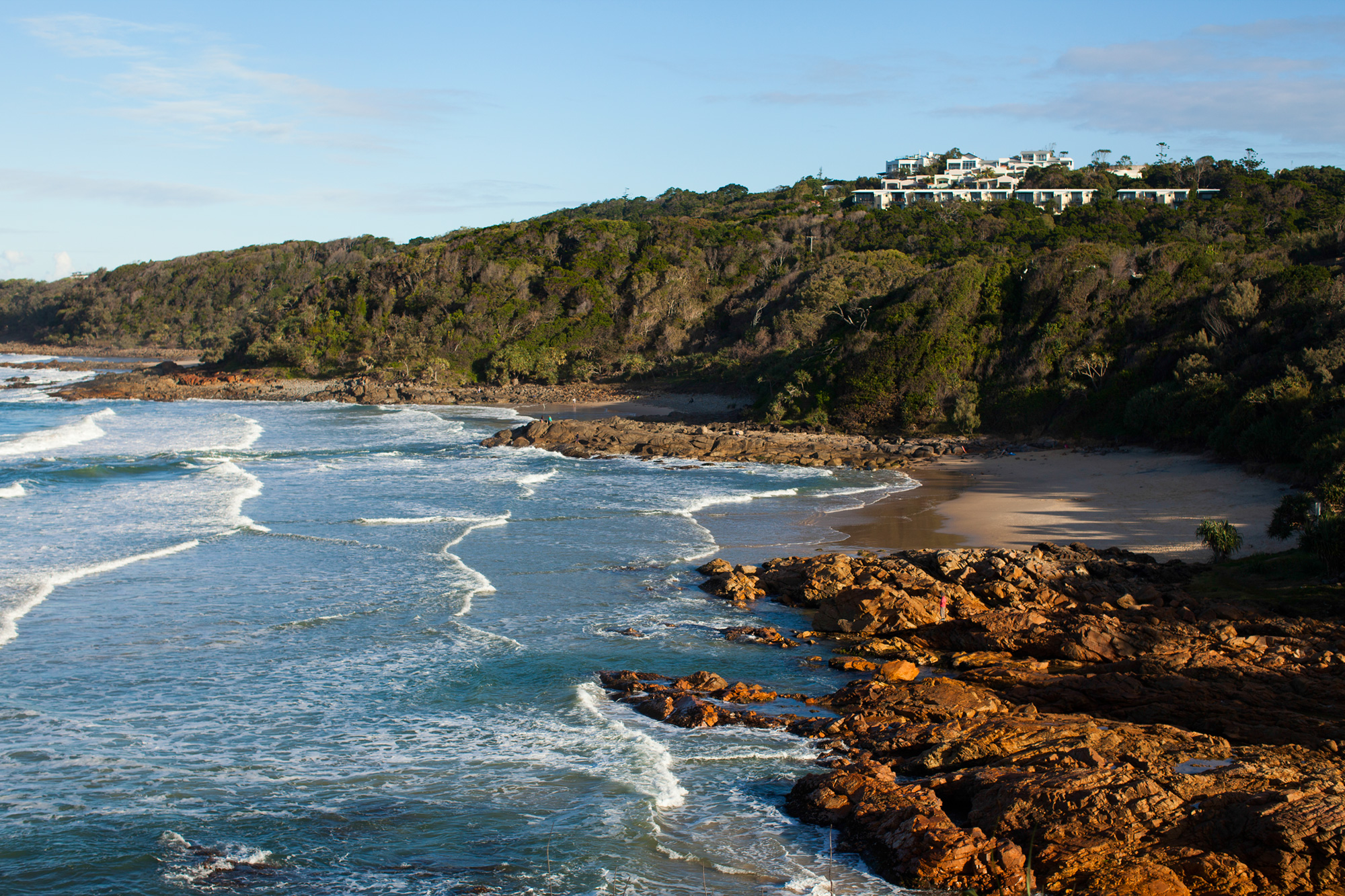 Waves roll onto a rocky, forested coastline with a hilltop building in the distance