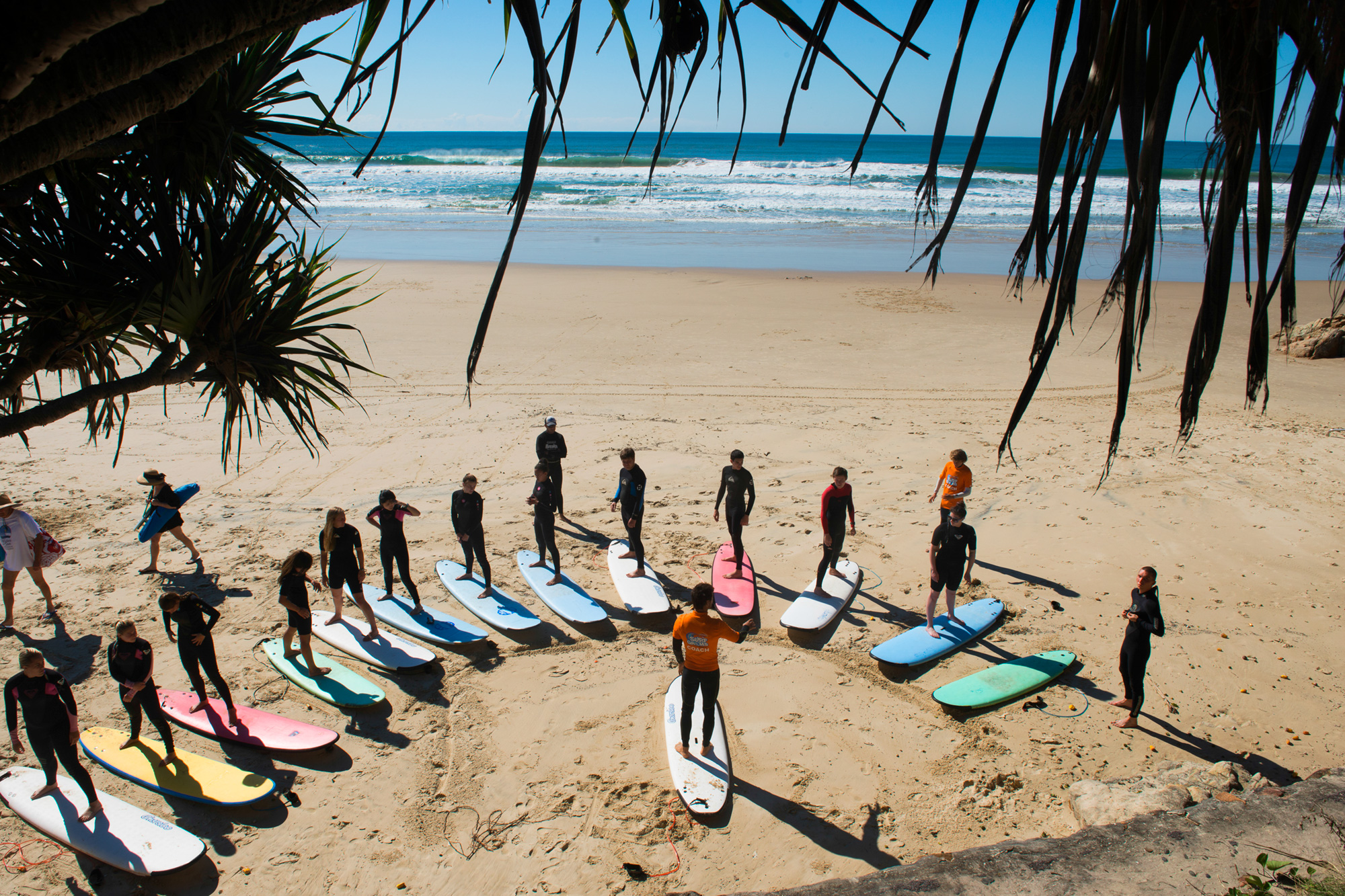 Surfboards laid out on the sand as students gather for a lesson under palms