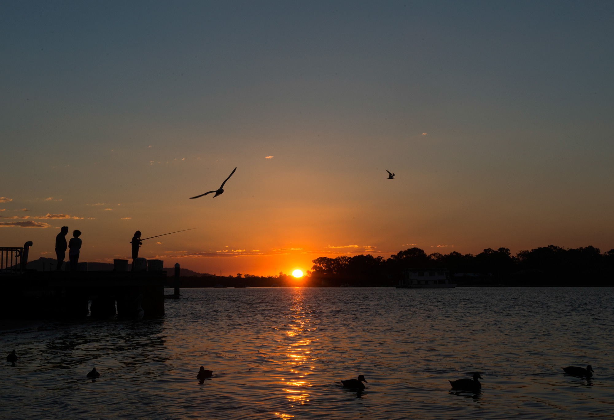 Teen travelers fish from a pier as birds fly over the water at sunset