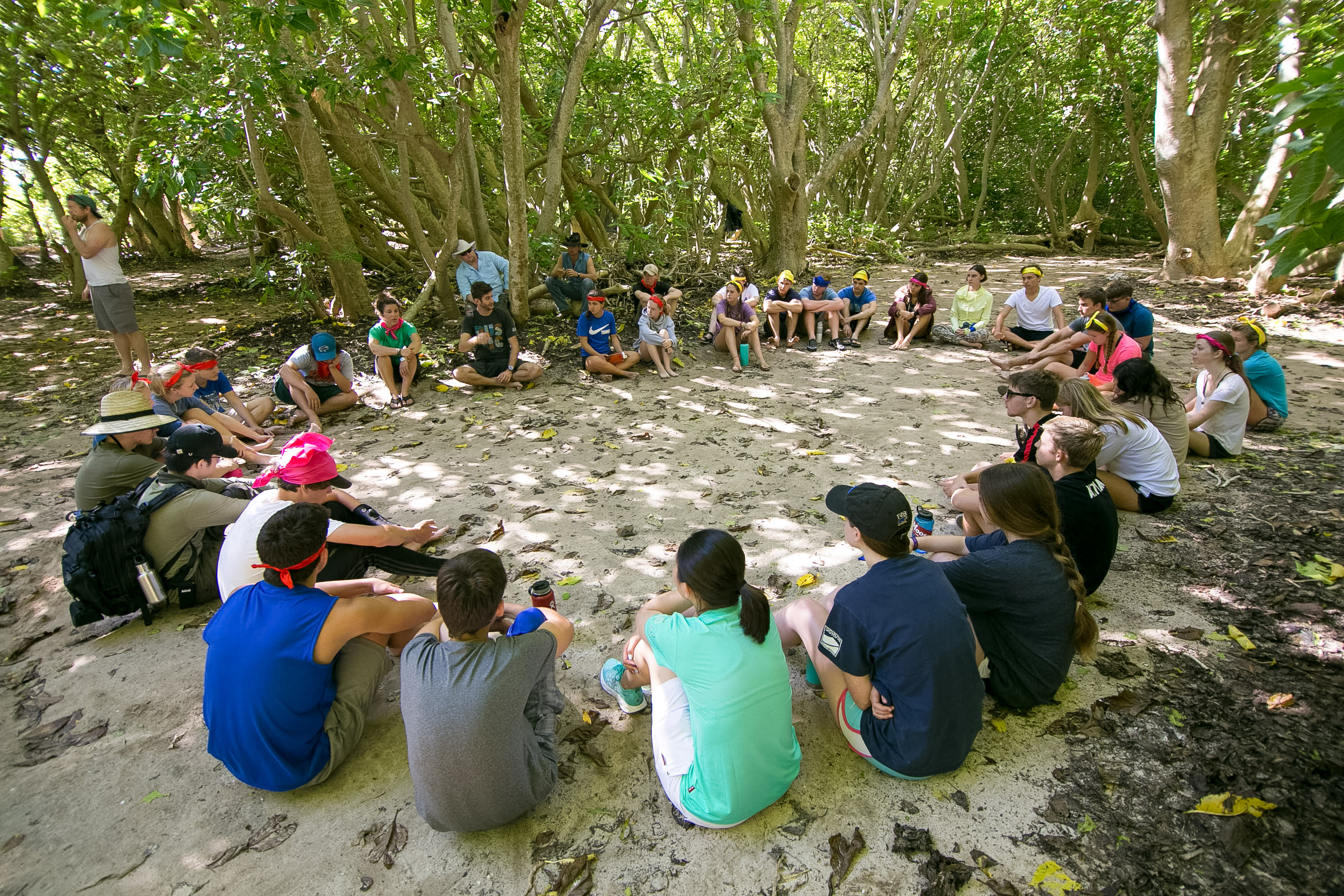 Students sit in a wide circle on open ground under trees for a group session