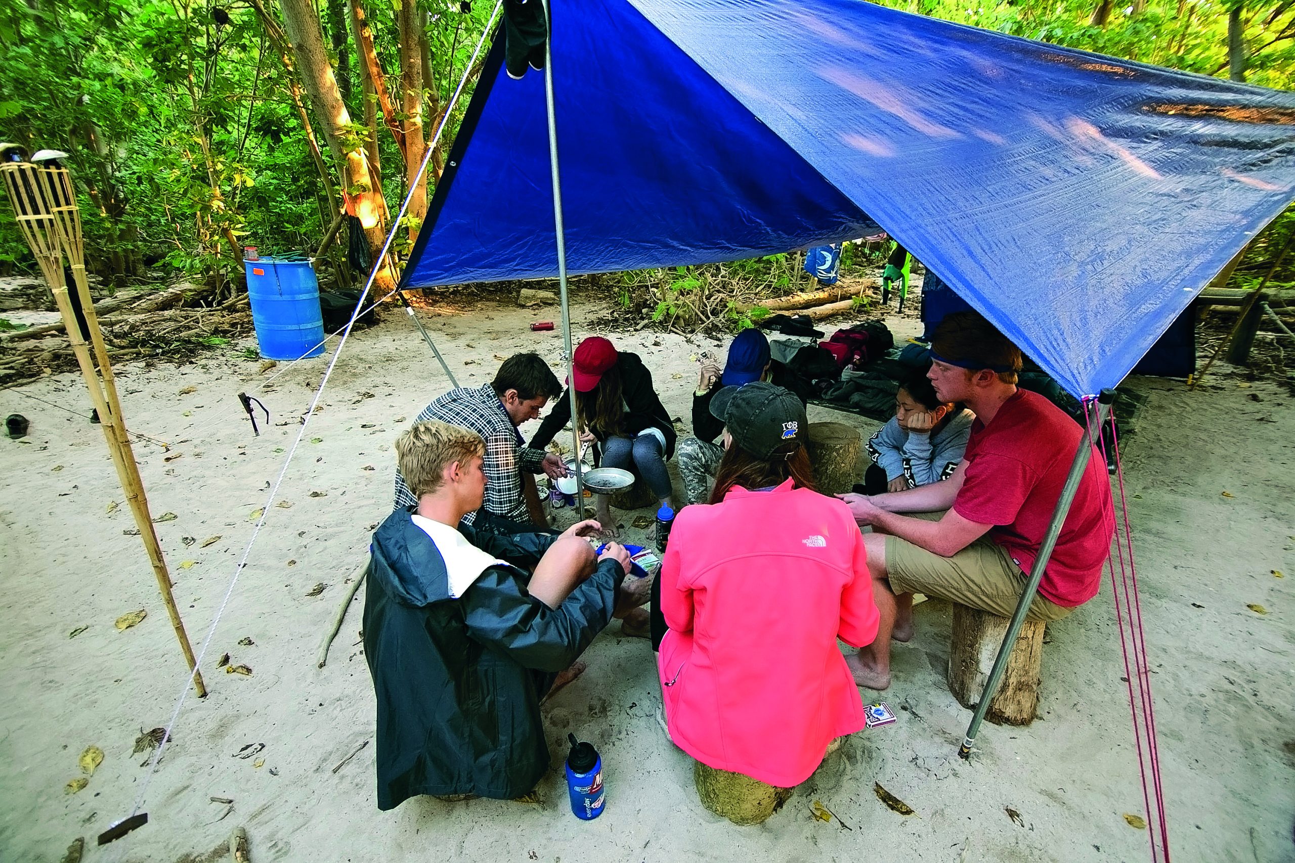 Students prepare food together under a blue tarp at a rainforest camp