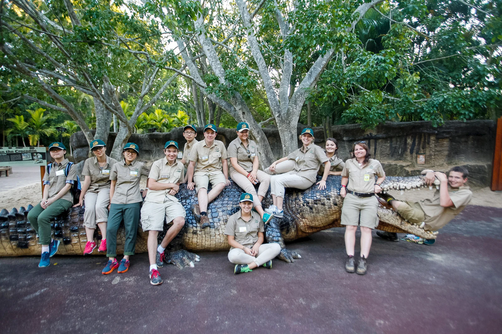 Students in khaki uniforms sit together along a giant crocodile statue under shady trees in Australia