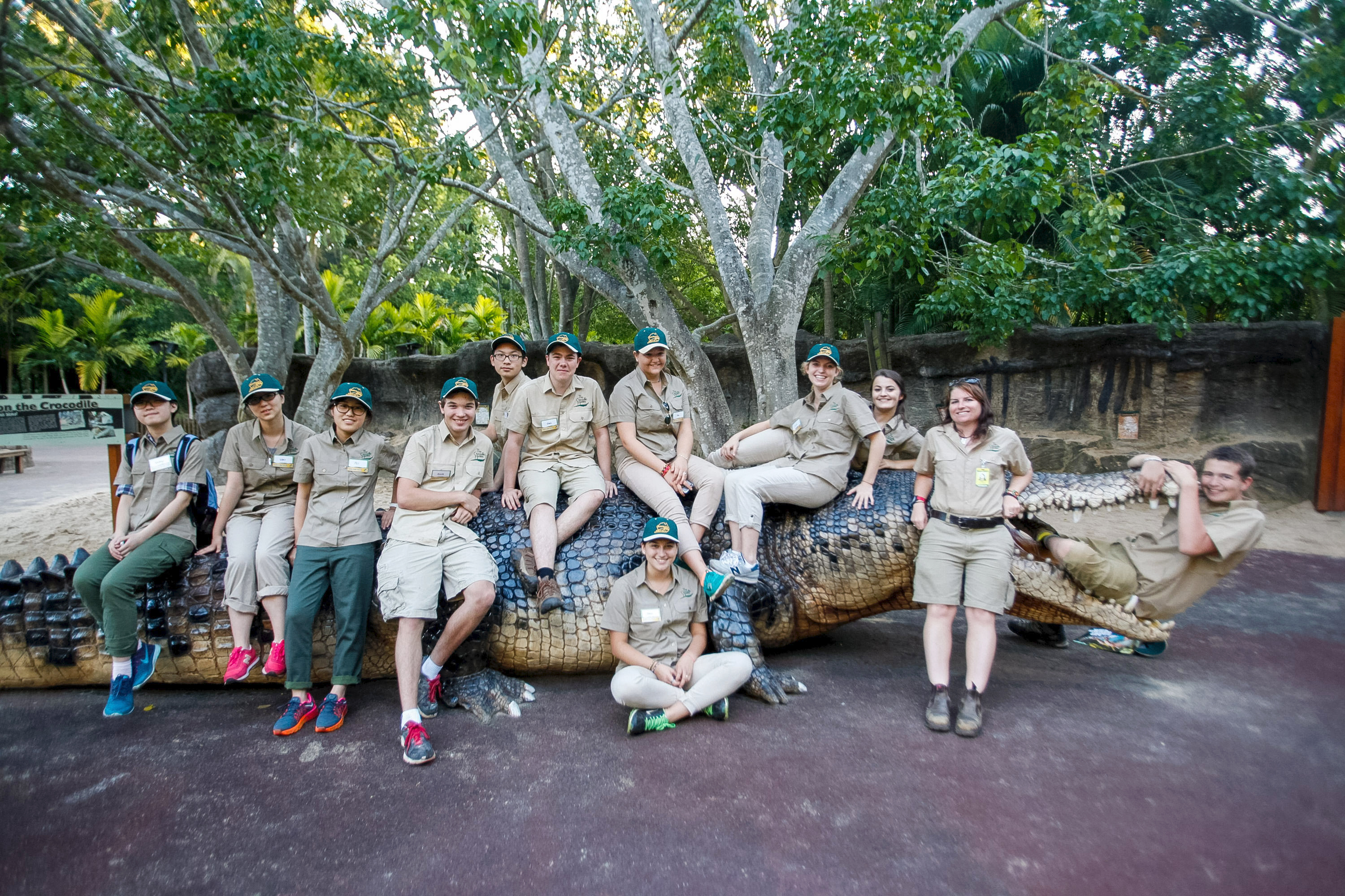 Students in zoo uniforms pose on a large crocodile statue among trees