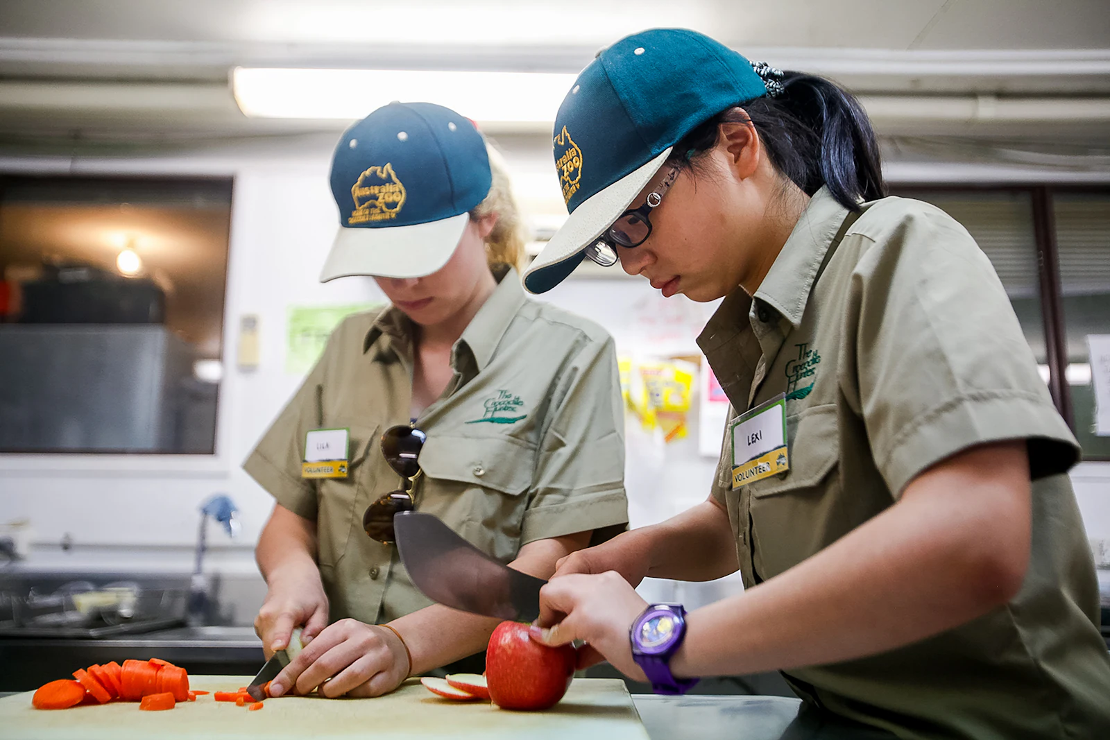 Two students in khaki uniforms and caps chop apples and carrots at a stainless steel kitchen bench