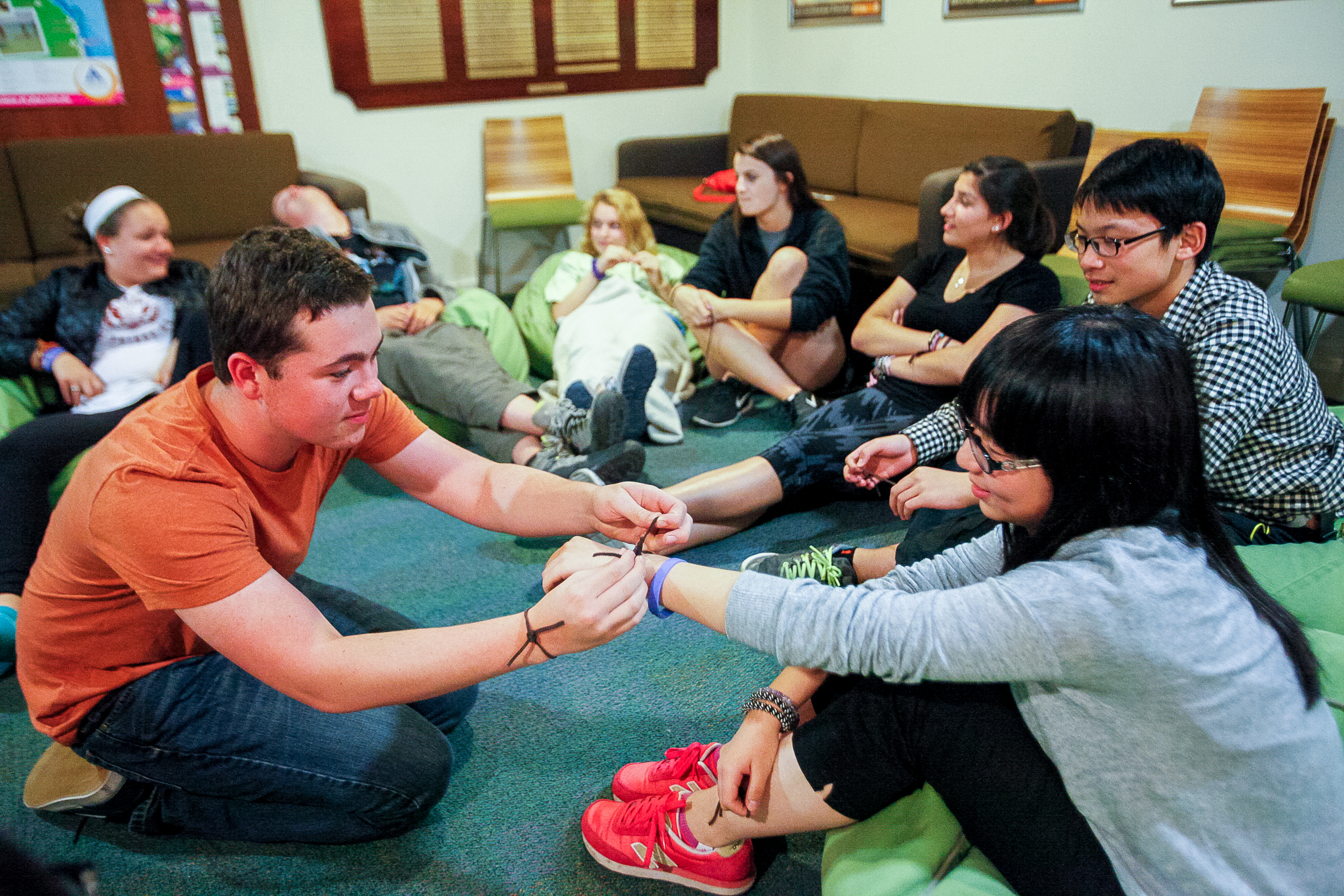 Students sit in a circle on a lounge floor tying friendship bracelets